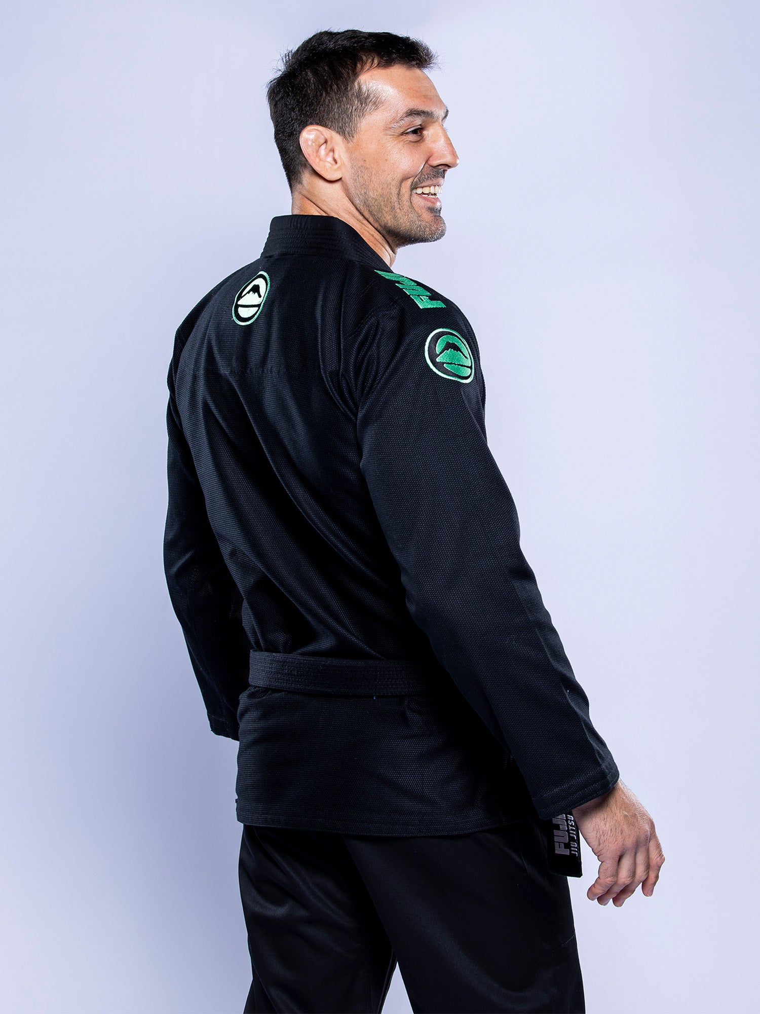 A man wears the Baseline 2.0 BJJ Gi Black Mint, featuring green and white patches on the sleeves and back, standing against a light gray background and smiling over his shoulder.