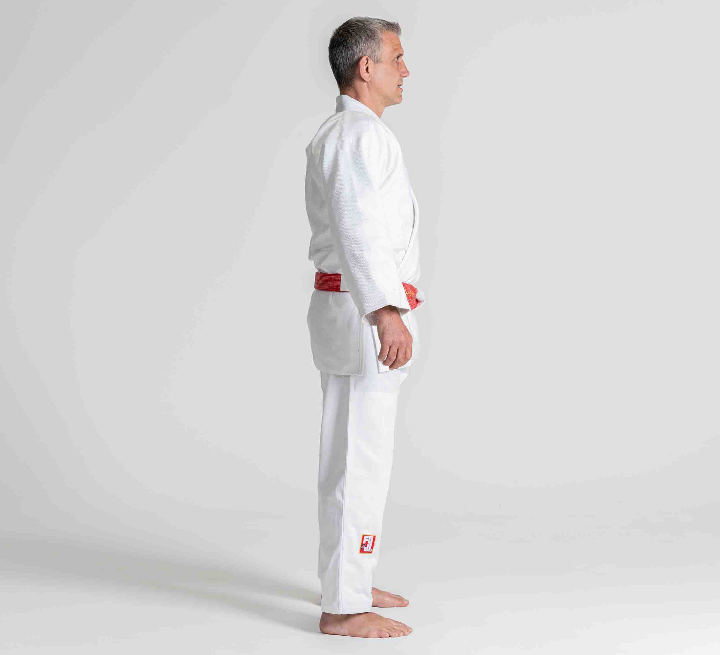 A person wearing the Euro Competition Judo Gi White with a red belt stands barefoot in a side profile against a plain white background.