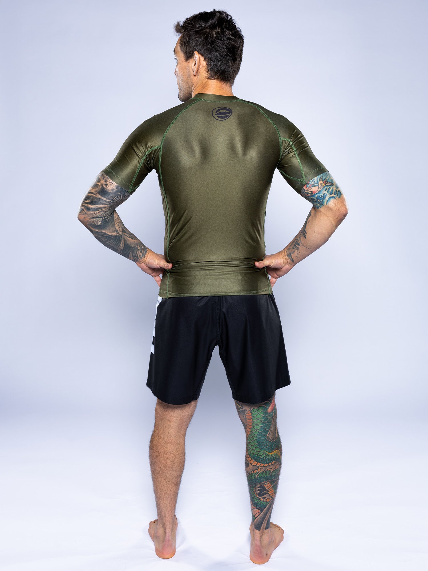 A man with tattoos stands barefoot on a white background, facing away, wearing the Baseline 2.0 Short Sleeve Rashguard in Military Green and black shorts, with his hands on his hips.