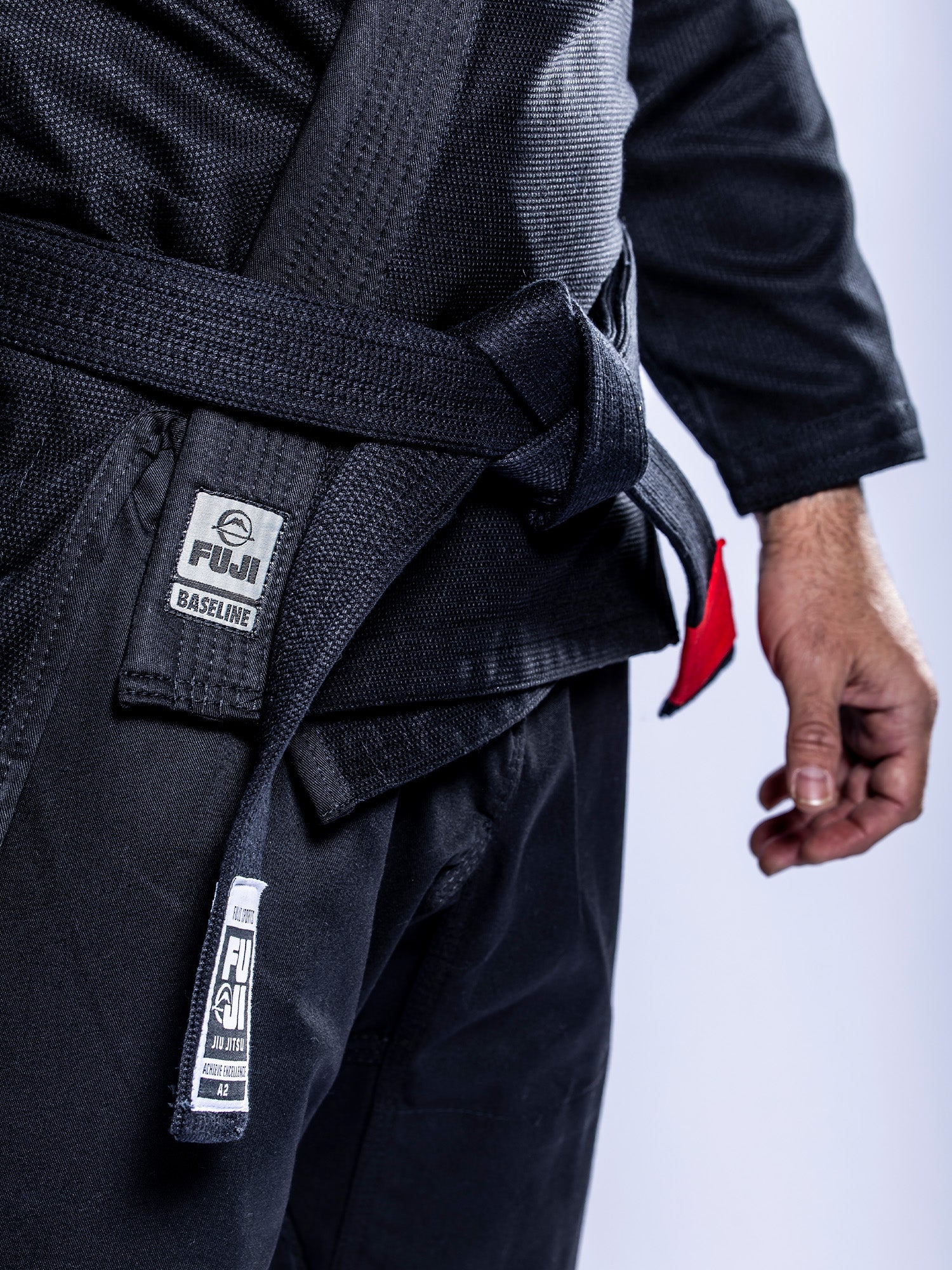A person stands against a light background wearing the Baseline 2.0 BJJ Gi Black Grey, featuring Fuji branding and a black belt, with their arm relaxed at their side.