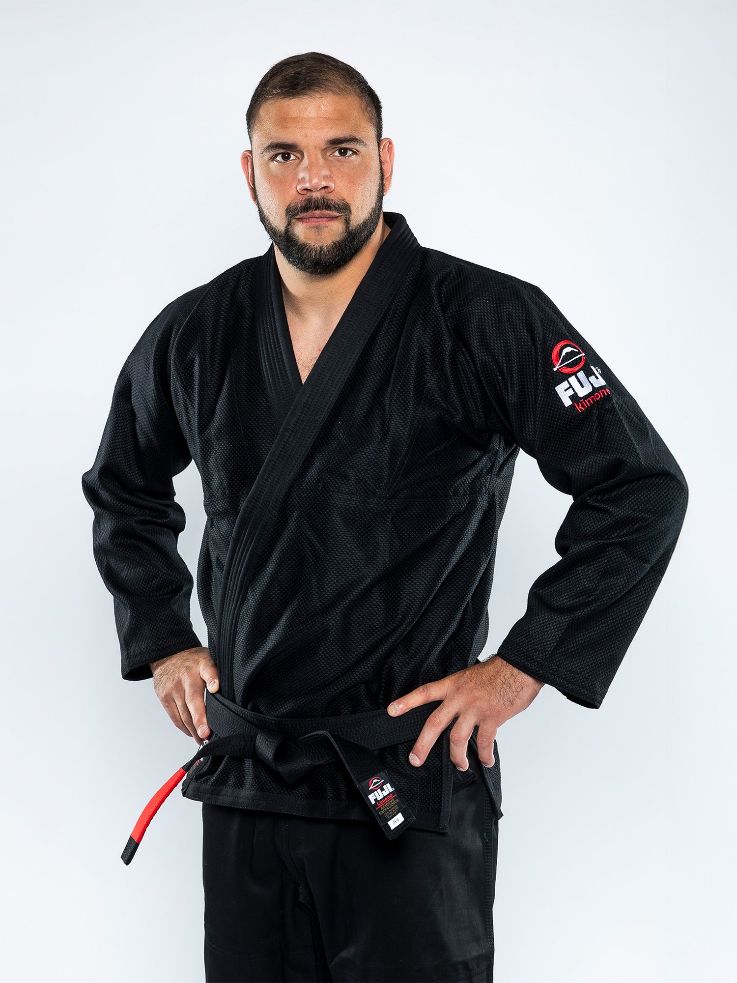 A bearded man wears the All Around BJJ Gi Black, featuring red and white patches. He stands against a plain white background, looking directly at the camera with his hands on his hips.