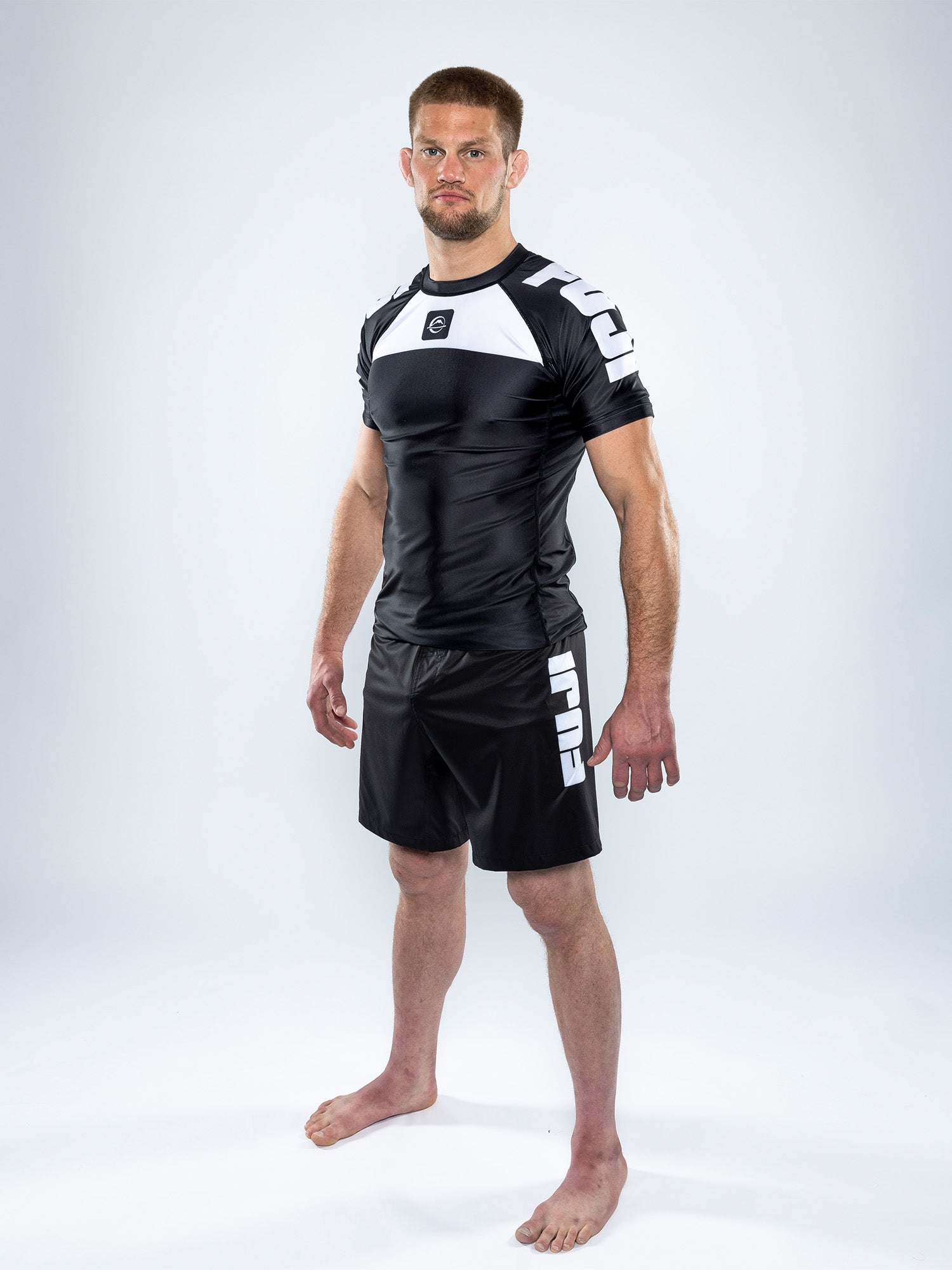 A man stands barefoot on a white background, wearing the Competition Ranked Rashguard Black—a black and white short-sleeve rash guard commonly worn for martial arts or grappling sports, paired with matching shorts.