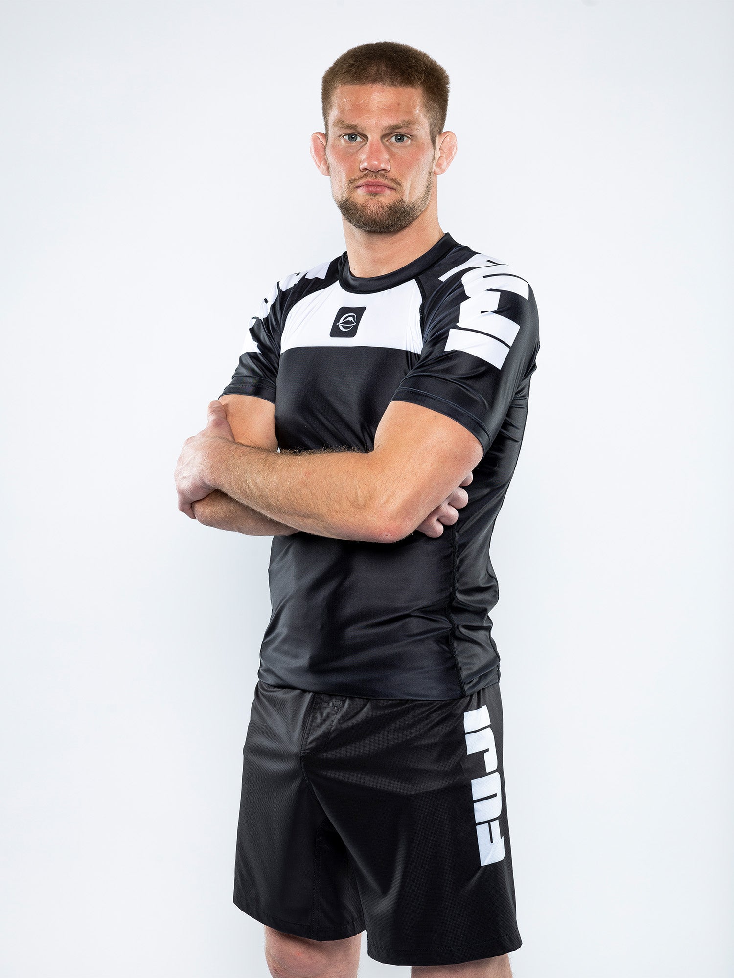 A man with short hair and a beard stands with arms crossed, wearing the Competition Ranked Rashguard Black and matching shorts with FUJI branding, against a plain white background.