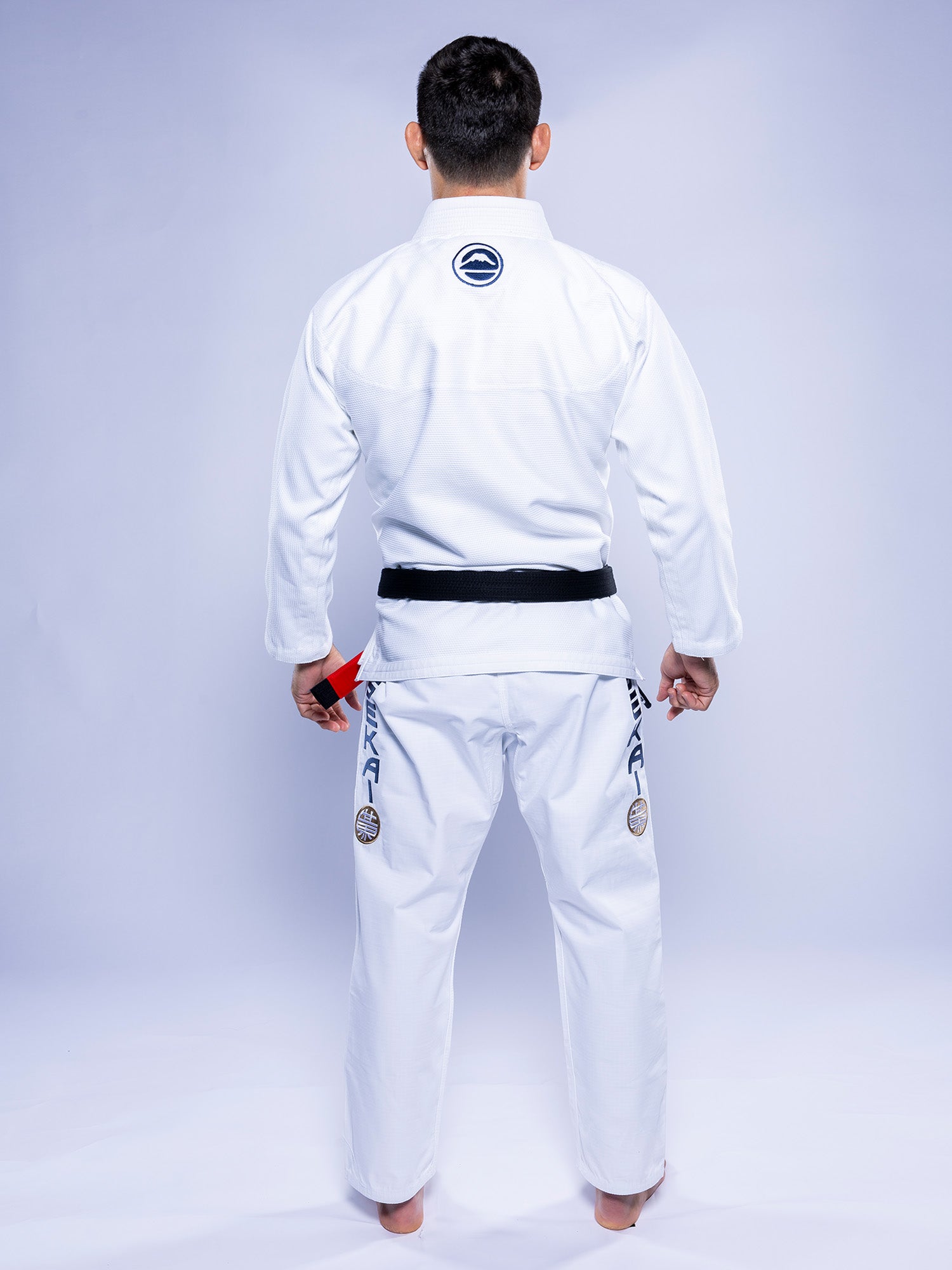 A person stands barefoot, facing away in the SEKAI 3.0 BJJ Gi White Navy with a black belt. The gi features patches on sleeves and pants, set against a plain light-colored background.