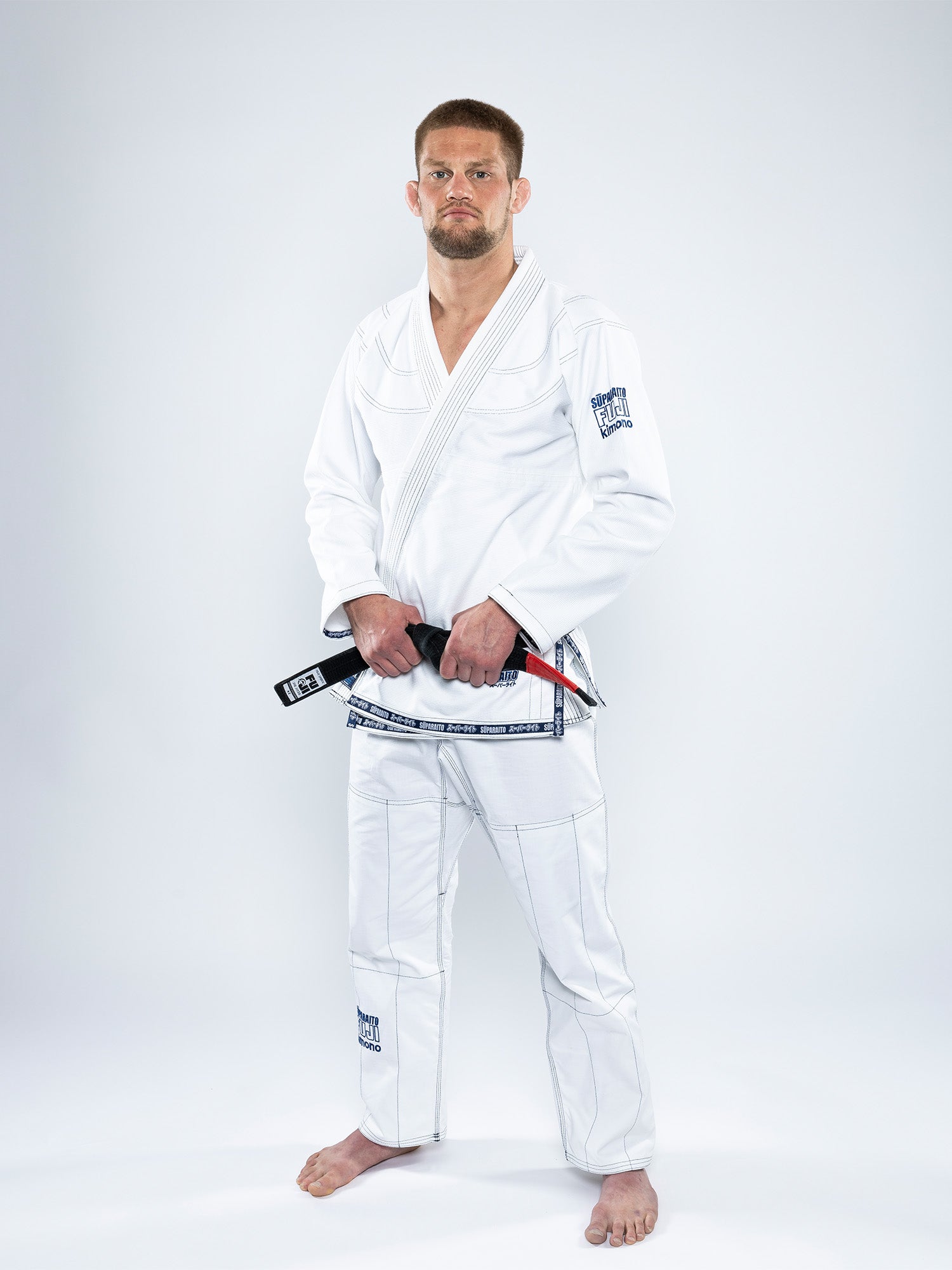 A man stands barefoot on a white background, wearing the Suparaito BJJ Gi White and holding a black belt with both hands, looking straight at the camera with a neutral expression.