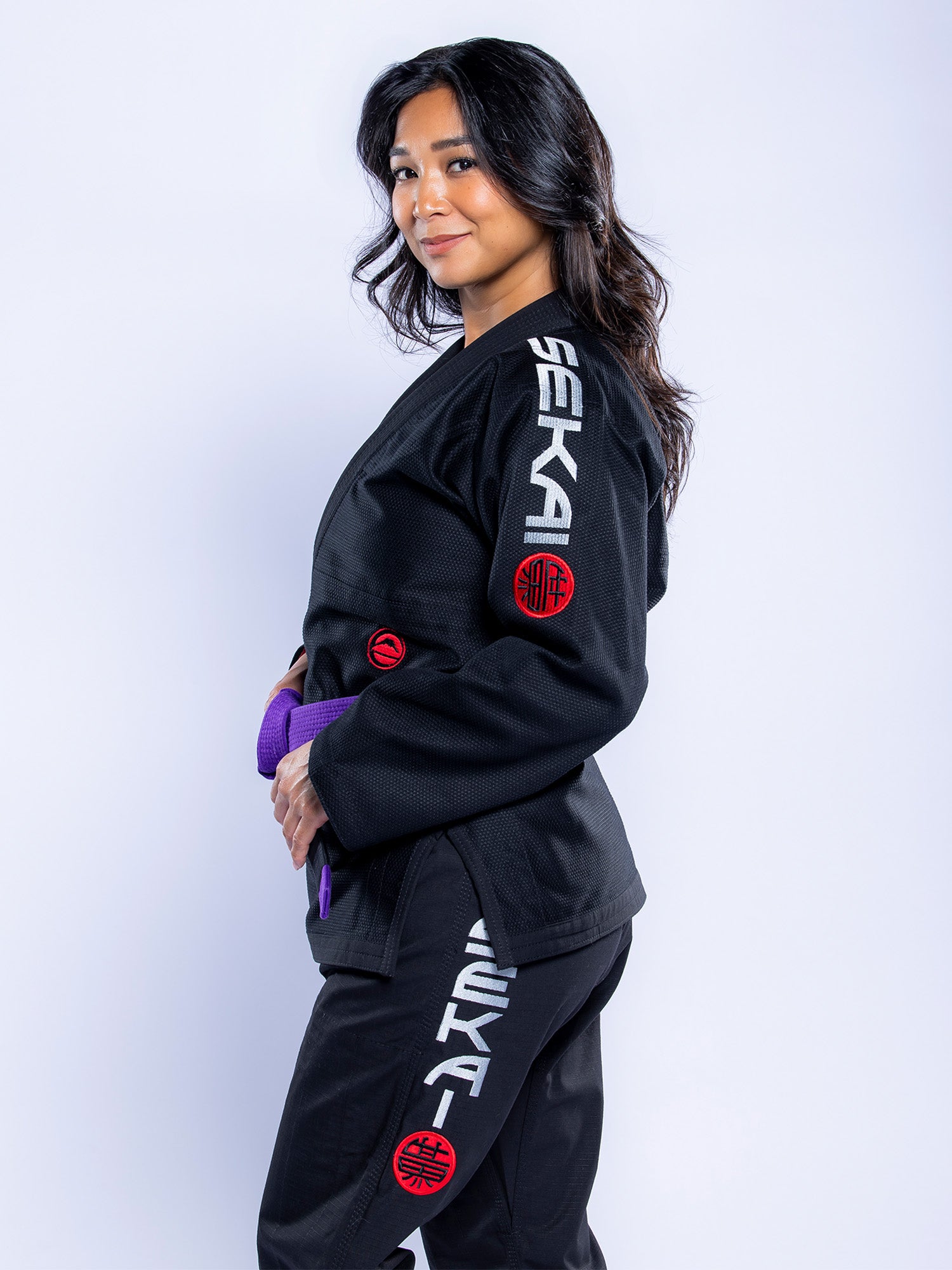 A woman with long dark hair wears the Womens SEKAI 3.0 BJJ Gi Black Grey, featuring SEKA on the sleeves and pants. She has a purple belt and smiles slightly at the camera against a light background.