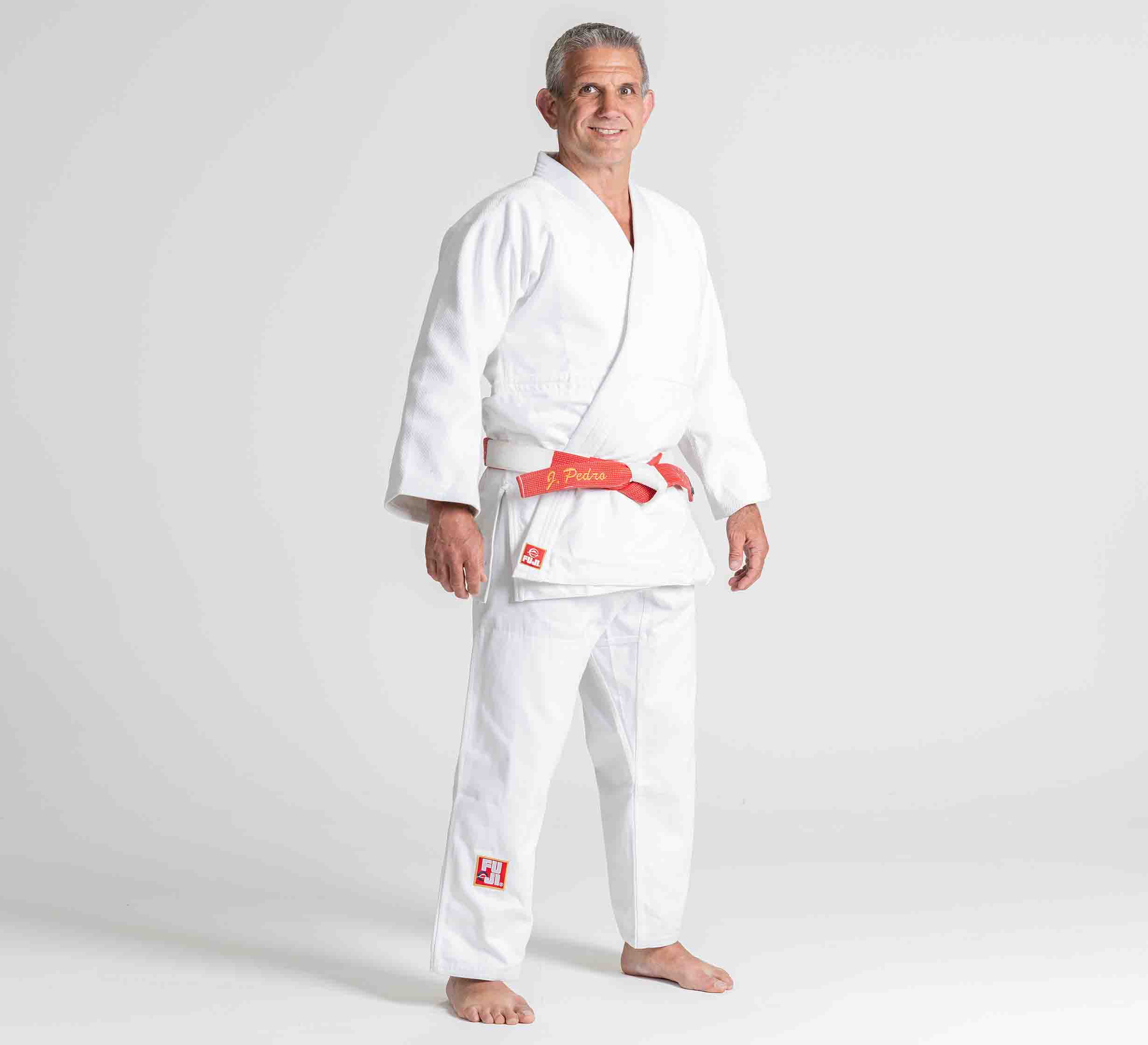A man wears the Euro Competition Judo Gi in white with a red and white belt, standing barefoot on a plain white background, smiling at the camera.