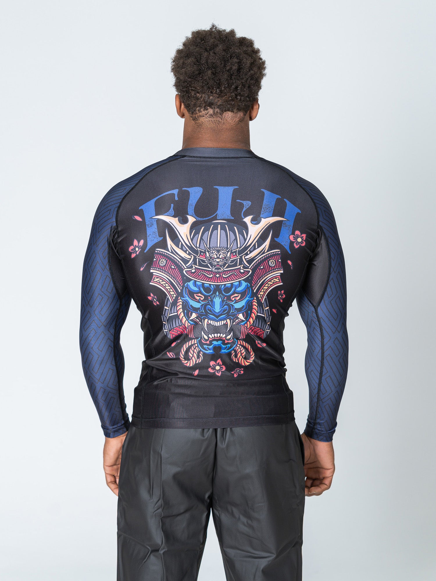Someone is seen from behind wearing the Oni Samurai Flex Lite Rashguard Blue, a long-sleeve black and blue athletic shirt with a large graphic of a snarling blue samurai tiger and "FUJI" across the upper back.