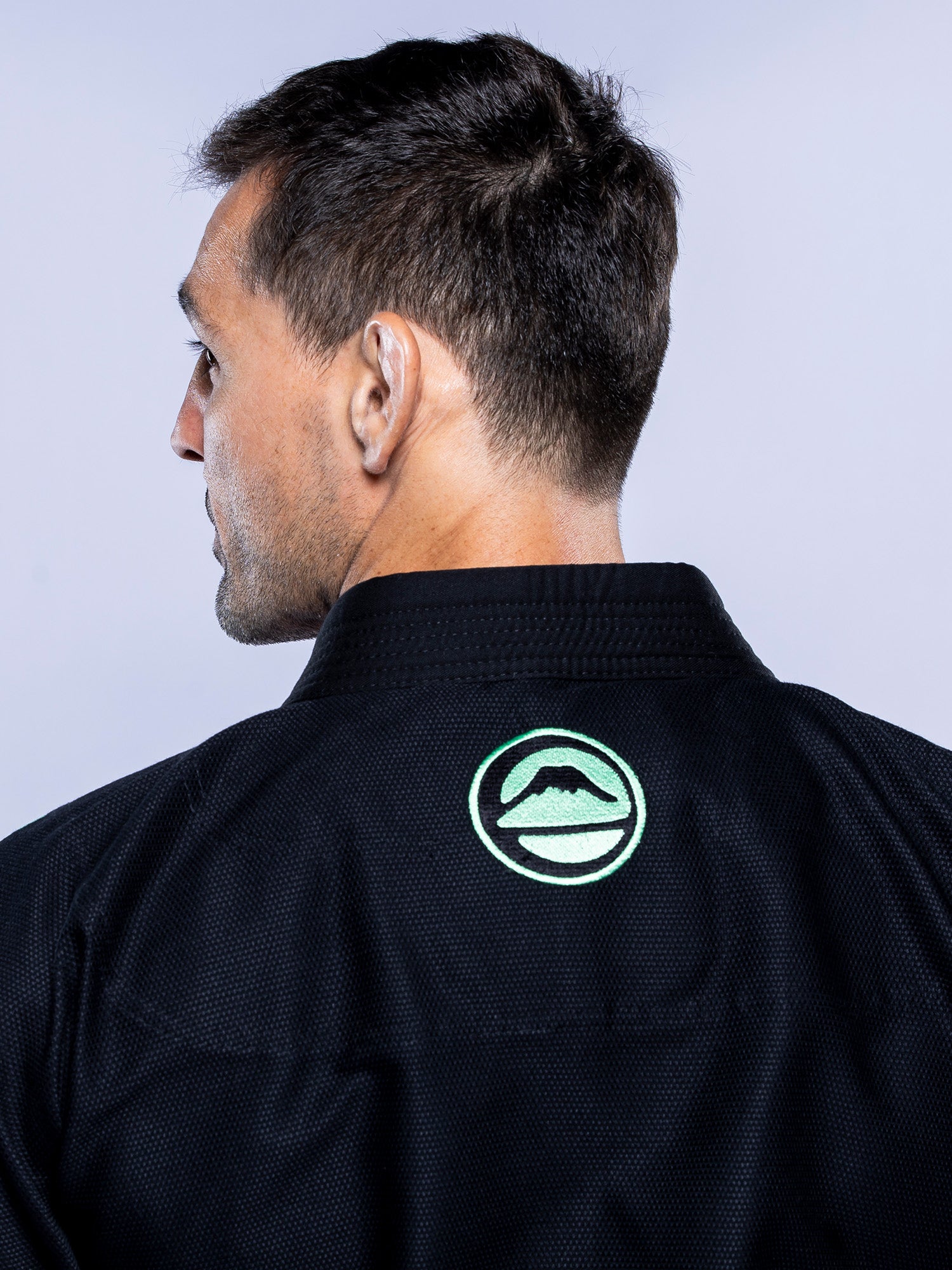 A man with short dark hair is seen from behind wearing the Baseline 2.0 BJJ Gi Black Mint, which features a circular mountain and wave logo on the back of the collar. The background is plain and light-colored.