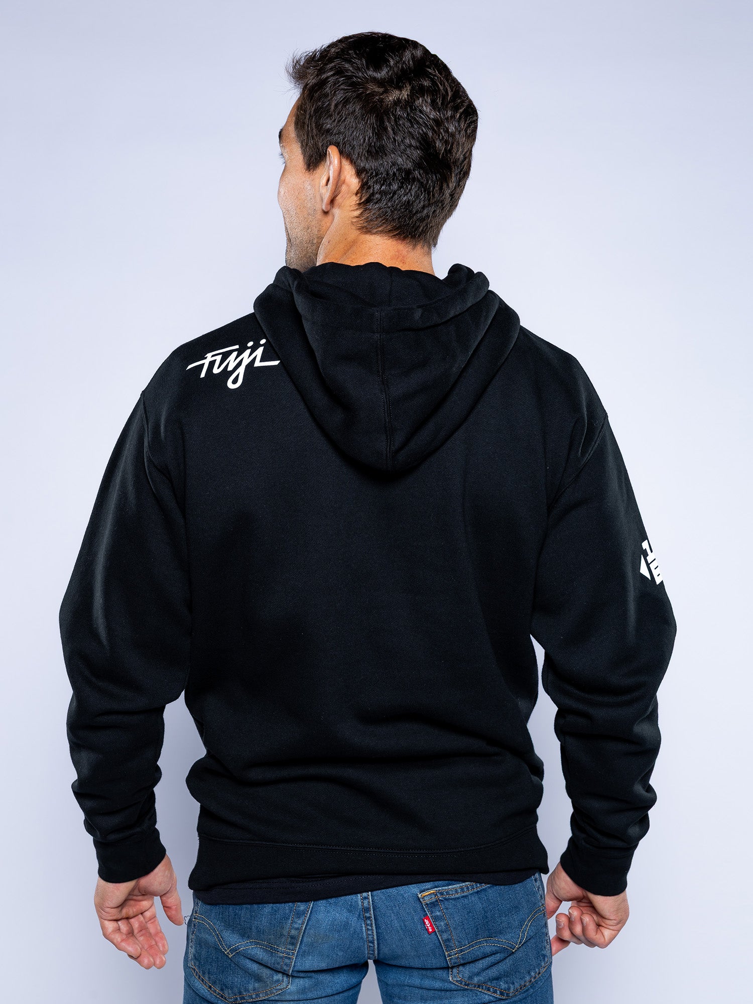 A man wearing the Midnight Collegiate Pull Over Hoodie Black—with "Fuji" on the right shoulder and a small white logo on the left sleeve—stands with his back to the camera, arms relaxed at his sides.