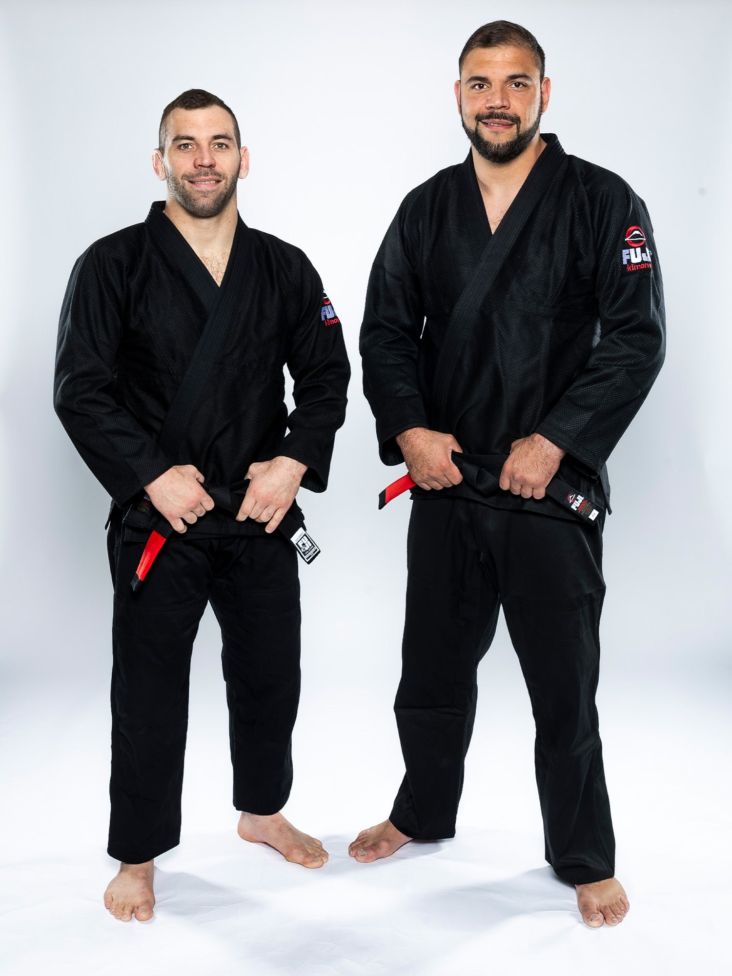 Two men wearing All Around BJJ Gi Black with black belts stand barefoot on a white background, smiling and posing for the camera.