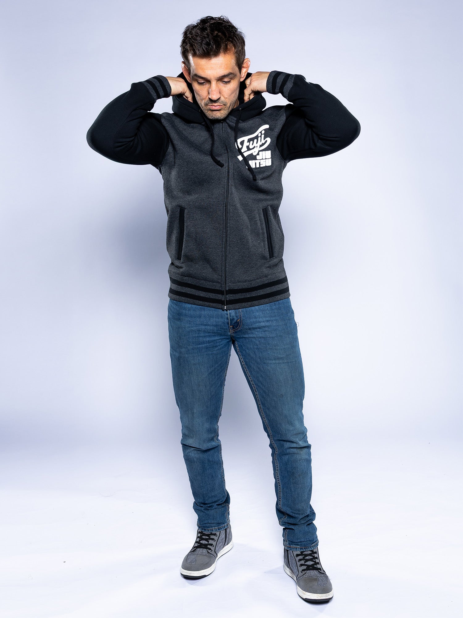 A man stands on a white background wearing the Jiu Jitsu Varsity Zip Hoodie Charcoal, blue jeans, and gray sneakers. He looks down with his hands touching the sides of his hood.