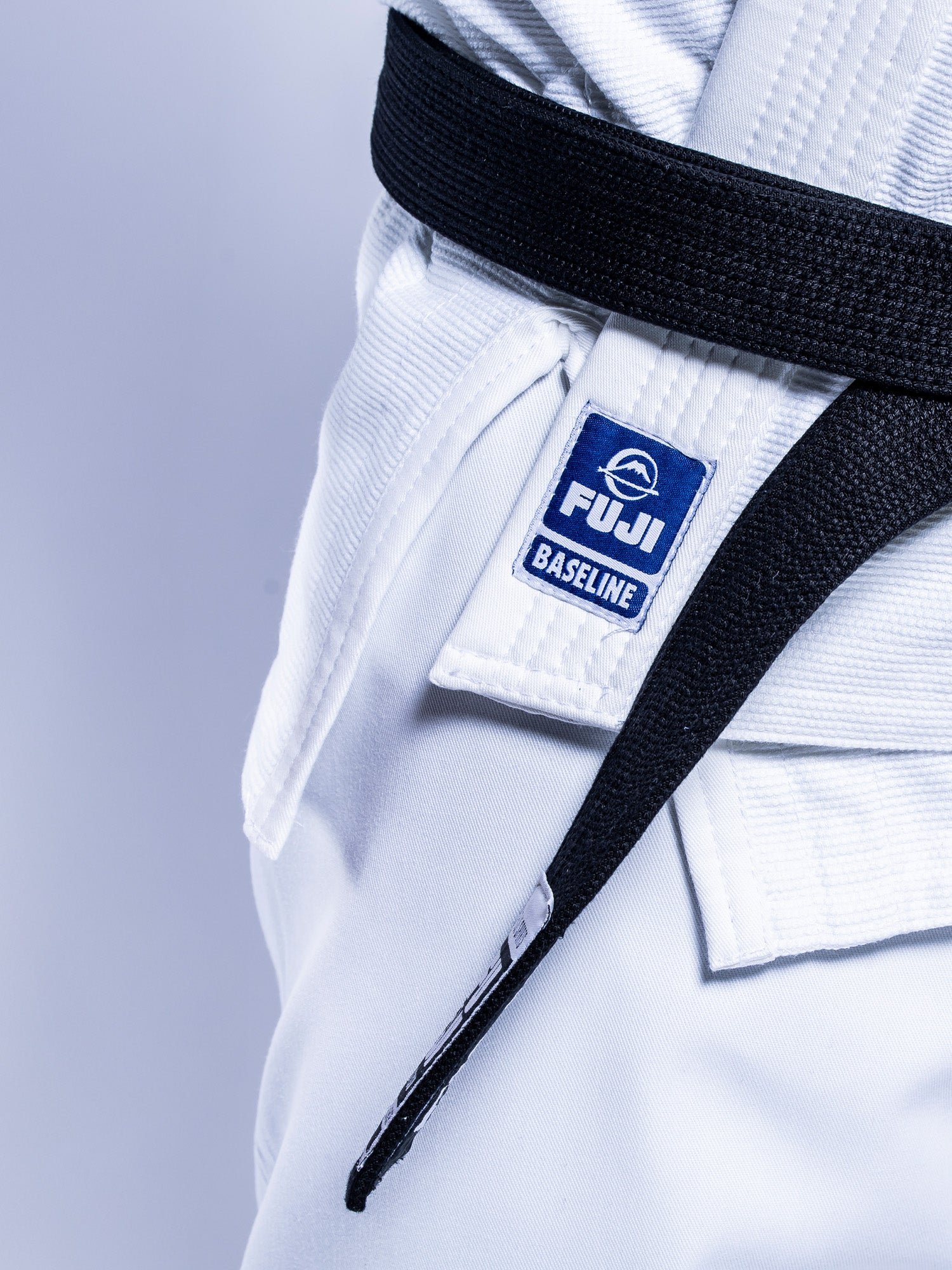 Close-up of a person wearing the Baseline 2.0 BJJ Gi White Navy, featuring a visible FUJI patch and a tied black belt, showing part of the torso and hip area.