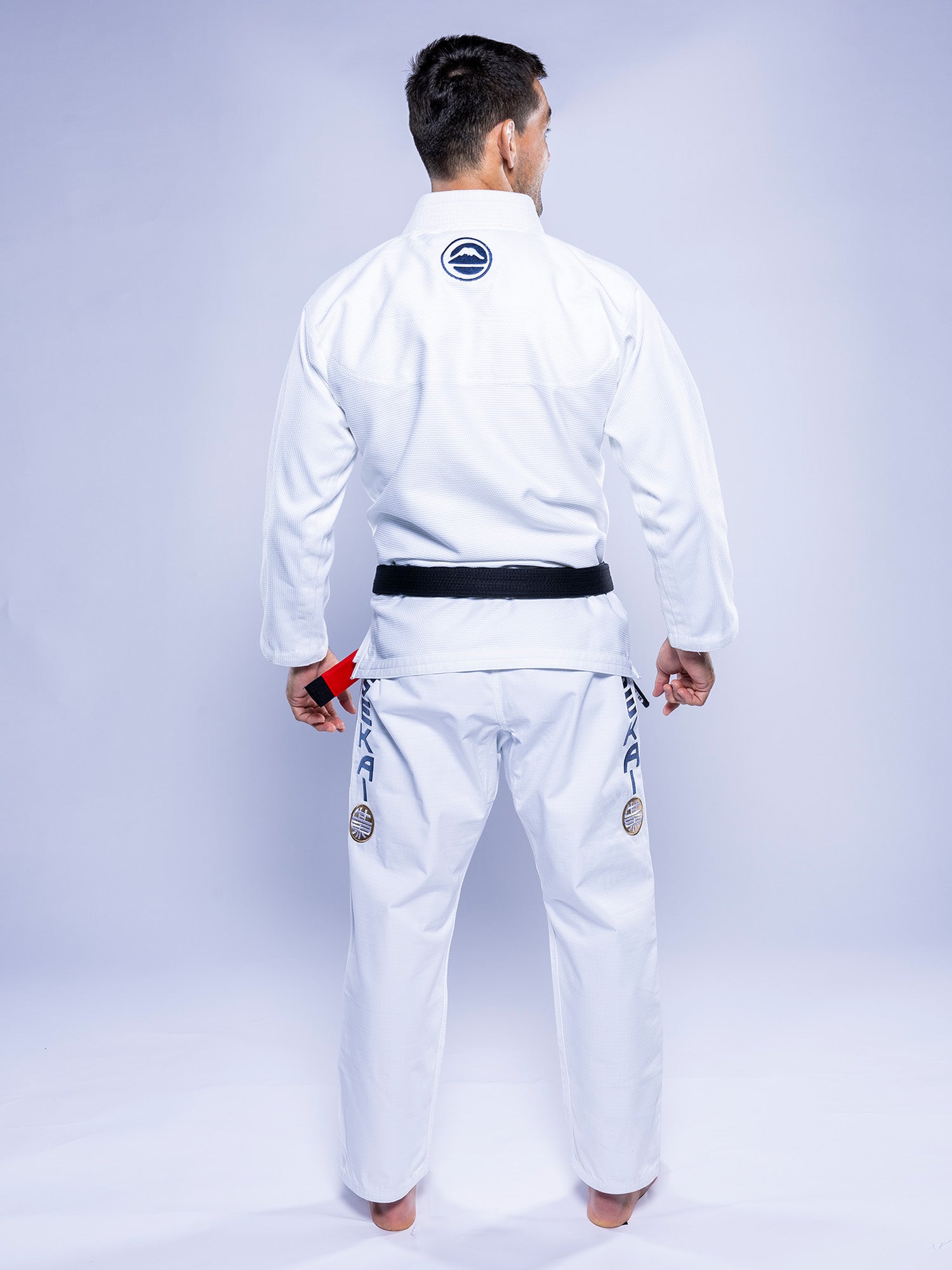 A man wearing a SEKAI 3.0 BJJ Gi White Navy with a black belt stands barefoot, facing away from the camera. The gi features patches on the back and legs, and he holds a red item in his left hand.