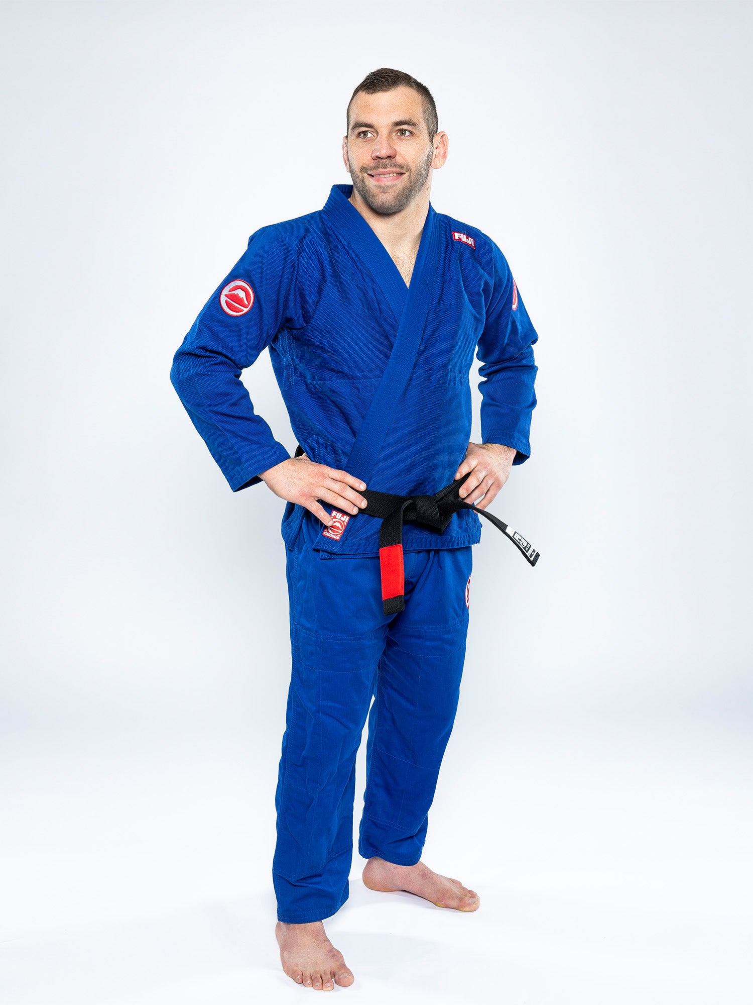 A man stands barefoot on a white background, smiling with his hands on his hips while wearing the Competition BJJ Gi Blue and a black belt.