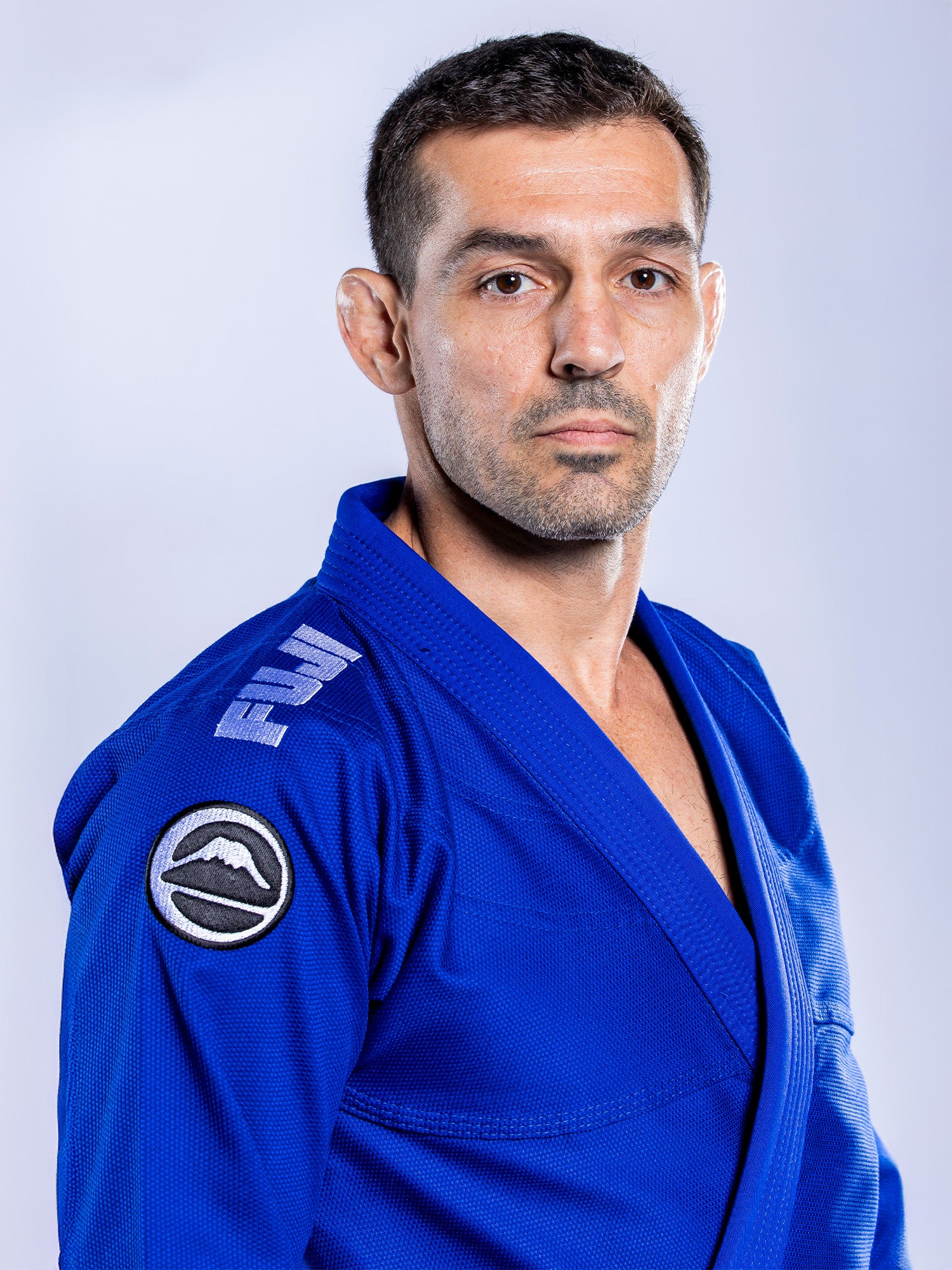A man with short dark hair and a trimmed beard models the Baseline 2.0 BJJ Gi Blue White, featuring a shoulder patch, as he stands against a plain light background and looks seriously at the camera.