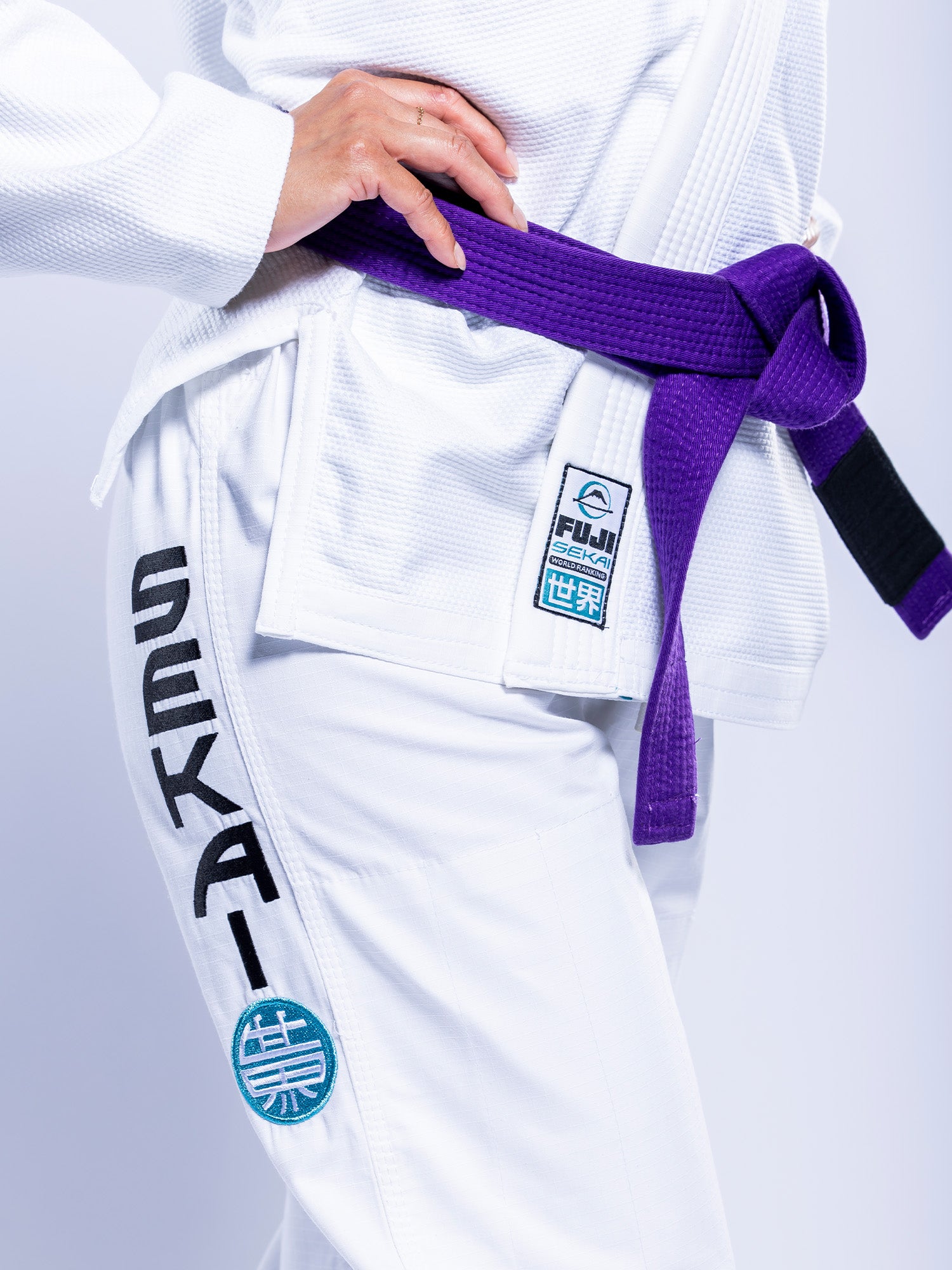 A person wears the Womens SEKAI 3.0 BJJ Gi White Teal with black SEKAI lettering and patches, a purple belt tied at the waist, and their hand resting on their hip.