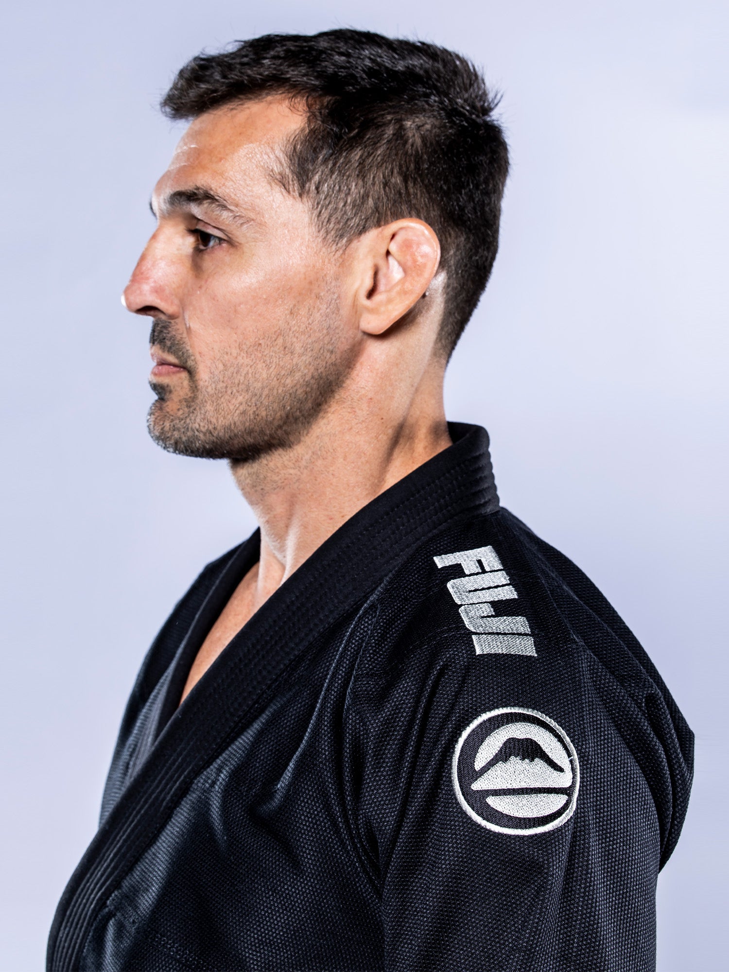 A man with short dark hair and stubble wears the Baseline 2.0 BJJ Gi Black Grey, featuring FUJI and a mountain logo on the shoulder, standing in profile against a light background.