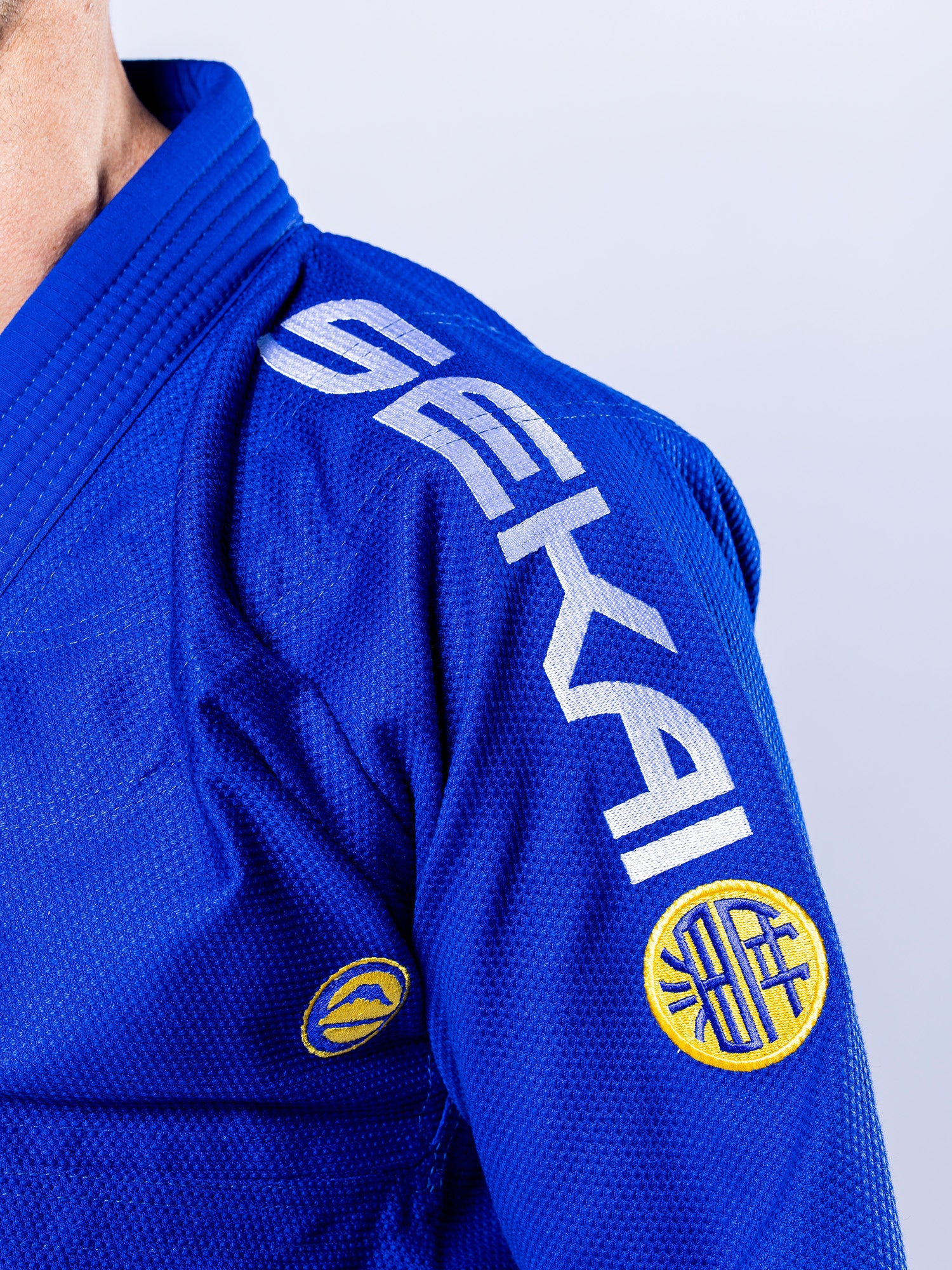 Close-up of a person wearing the SEKAI 3.0 BJJ Gi Blue Yellow, featuring "SEKAI" in white on the shoulder and two yellow circular patches on the upper arm against a plain light background.