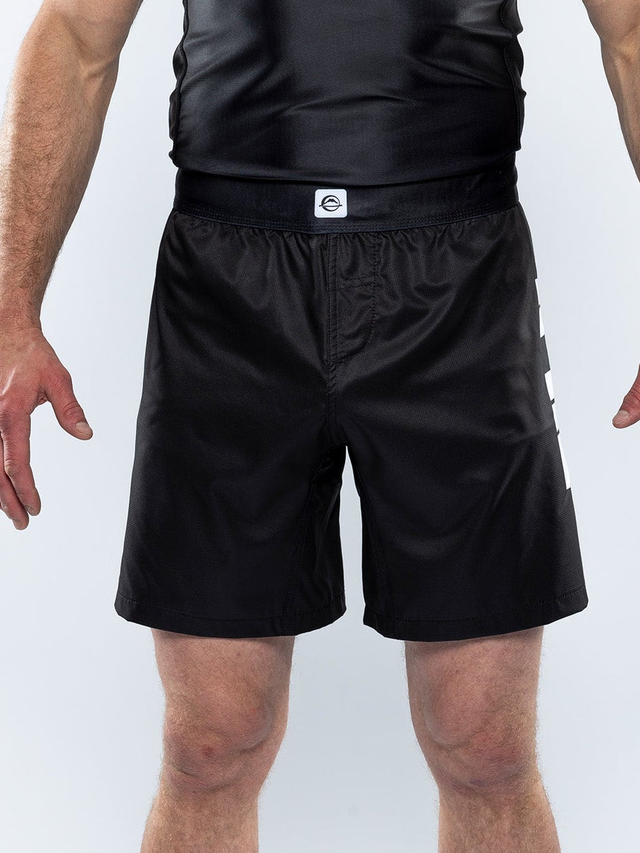 A person wears Kids Competition Ranked Shorts Black, featuring white lettering on the side, and stands with arms slightly outstretched against a plain light background.
