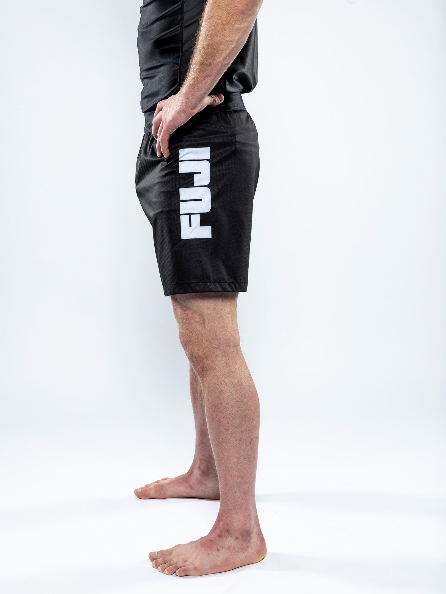 A person stands barefoot on a white surface, viewed from the side, wearing Competition Ranked Shorts Black with "FUJI" printed in white on the left leg.