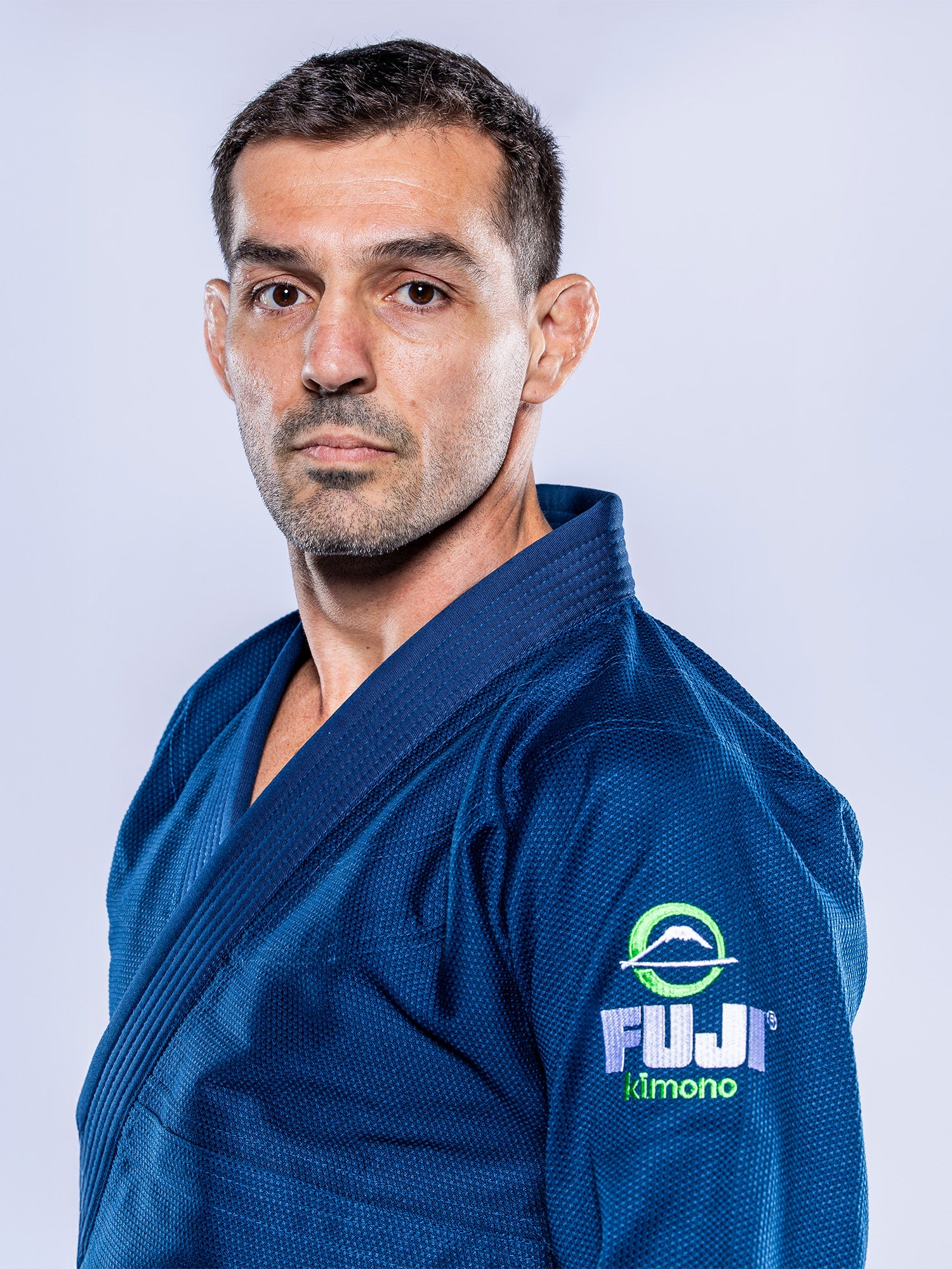 A man with short hair and a serious expression stands facing forward against a light gray background, wearing the Lightweight BJJ Gi Navy, which features a circular FUI Kimono logo on the sleeve.