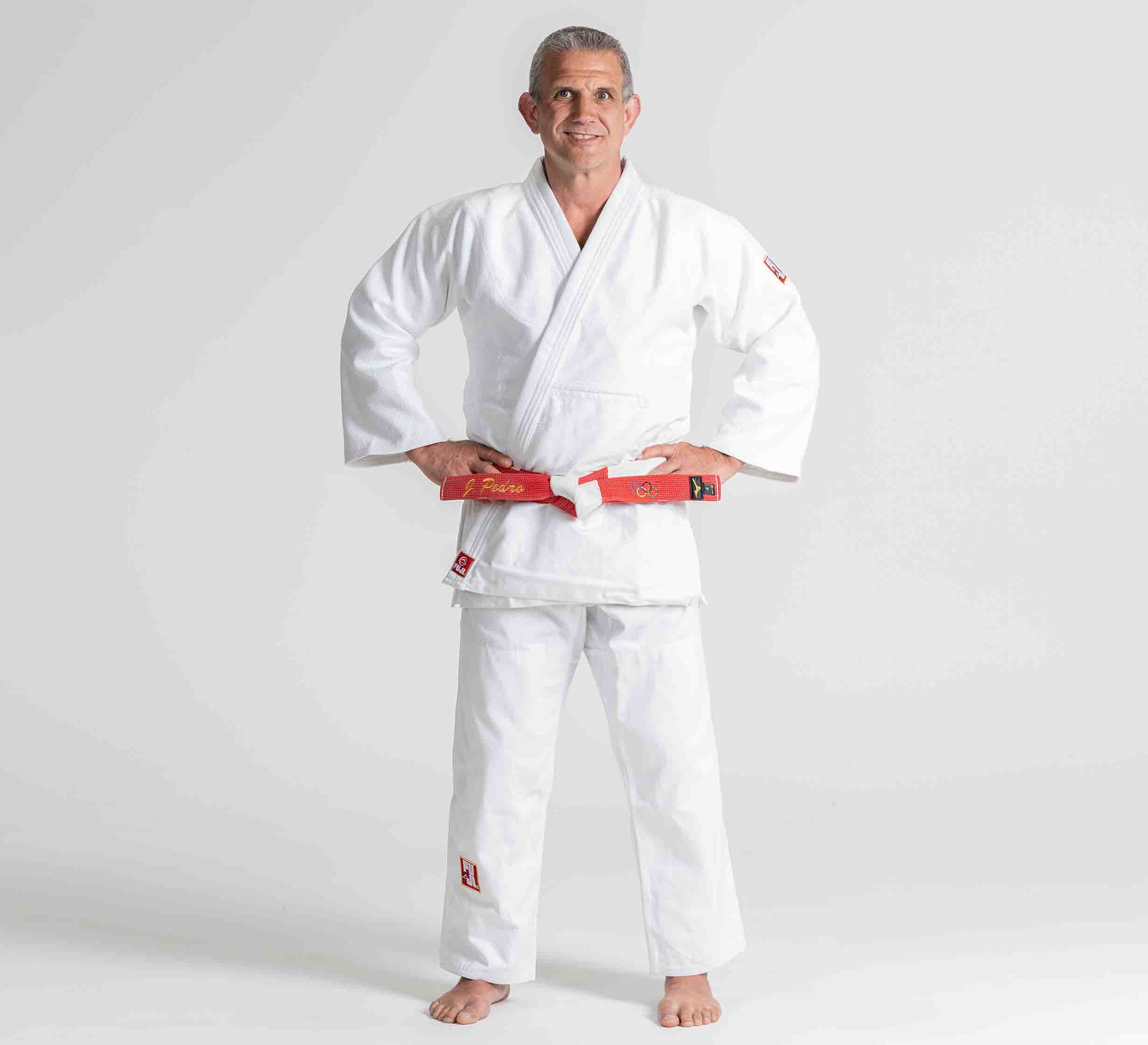 A man stands barefoot on a white background, smiling with his hands on his hips. He is wearing the Euro Competition Judo Gi White and a red belt.