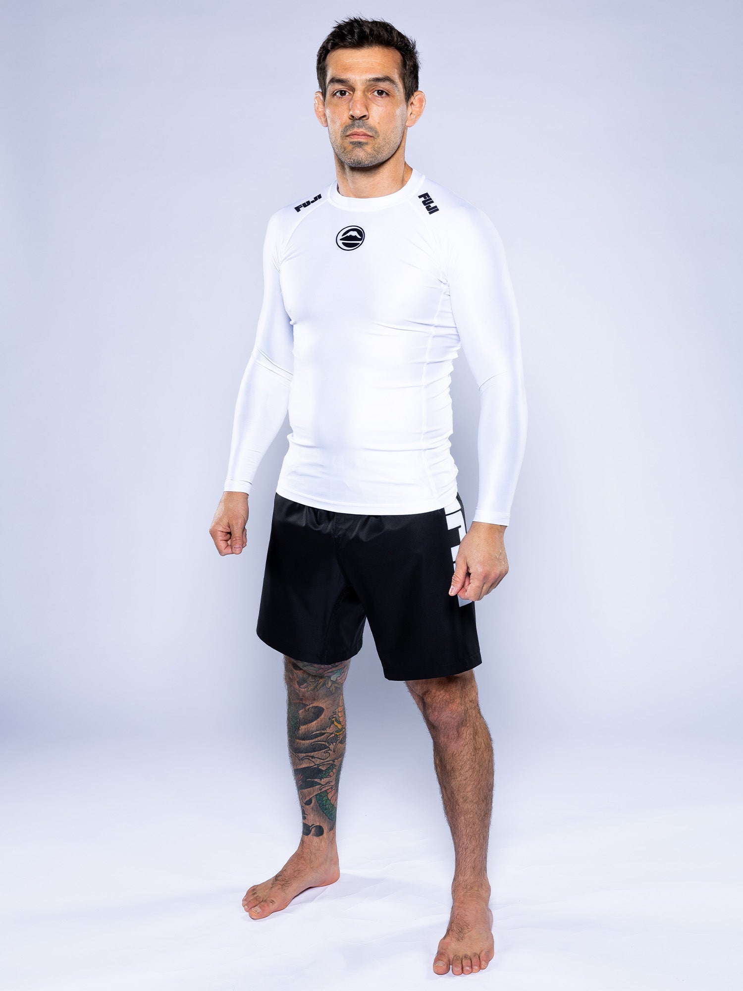A man with short dark hair and a tattoo on his left leg stands barefoot on a white background, wearing the Baseline 2.0 Long Sleeve Rashguard in white and black athletic shorts.