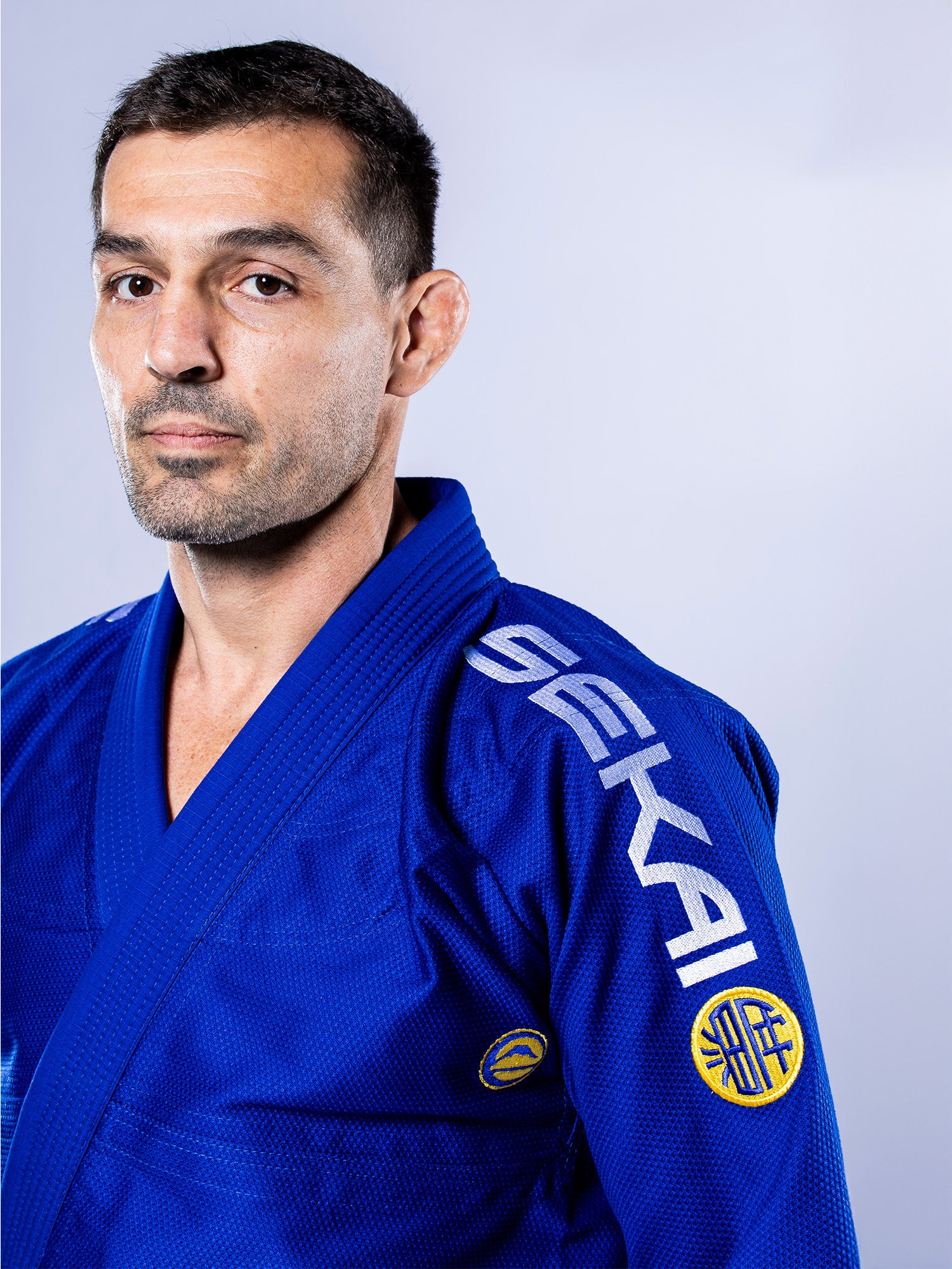 A man models the SEKAI 3.0 BJJ Gi Blue Yellow, featuring SEKAI branding on the shoulder, as he stands against a light background and looks seriously at the camera.