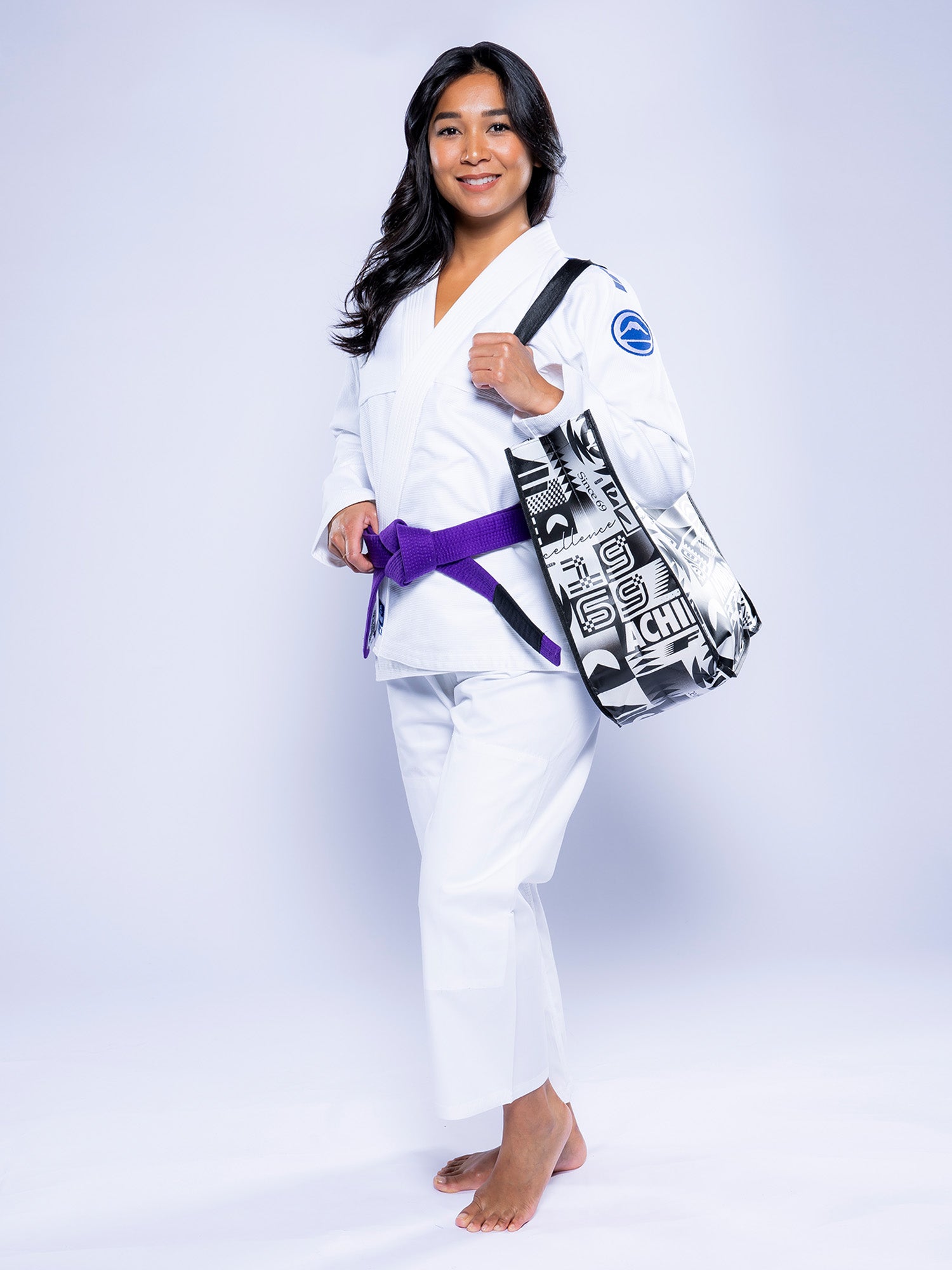 A woman wearing the Womens Baseline 2.0 BJJ Gi White Navy with a purple belt stands barefoot, smiling and holding a black-and-white patterned tote bag over her shoulder against a plain light background.