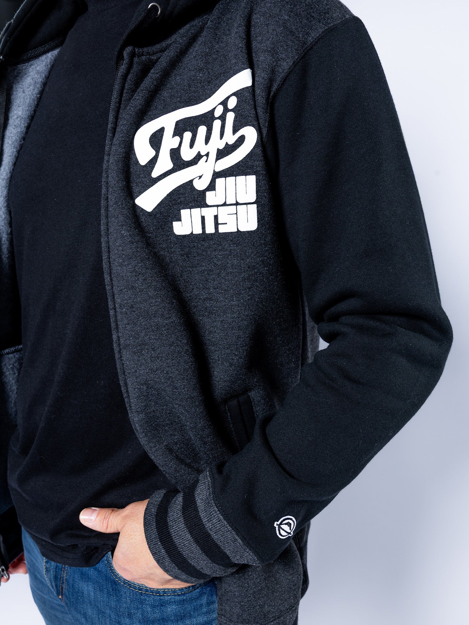 A person wears the Jiu Jitsu Varsity Zip Hoodie Charcoal, featuring "Fuji Jiu Jitsu" in white on the chest and a small logo near the cuff, with one hand tucked into their jeans pocket.
