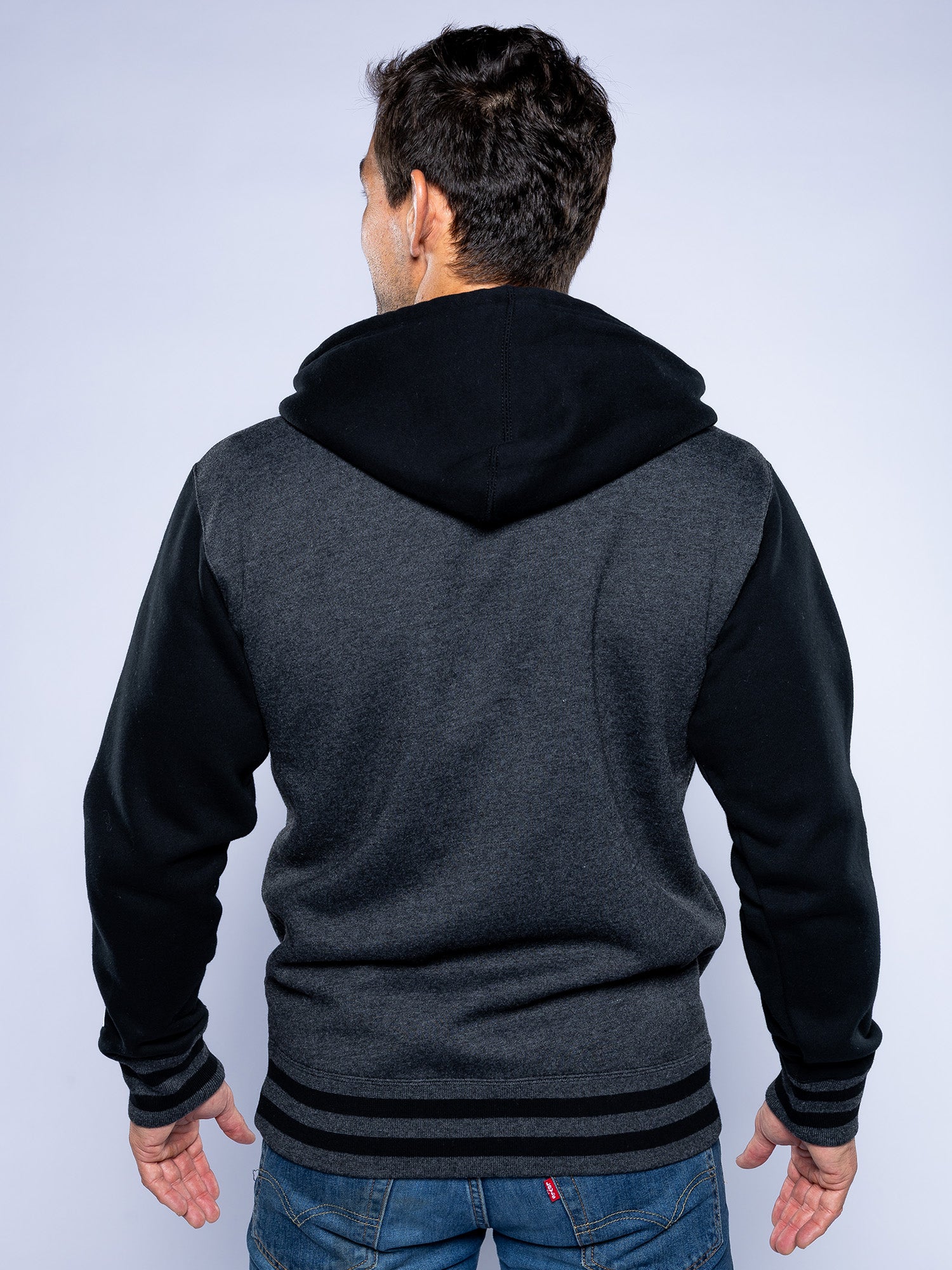 A man faces away from the camera wearing the Jiu Jitsu Varsity Zip Hoodie Charcoal with black sleeves and hood, paired with blue jeans, standing against a plain light background.