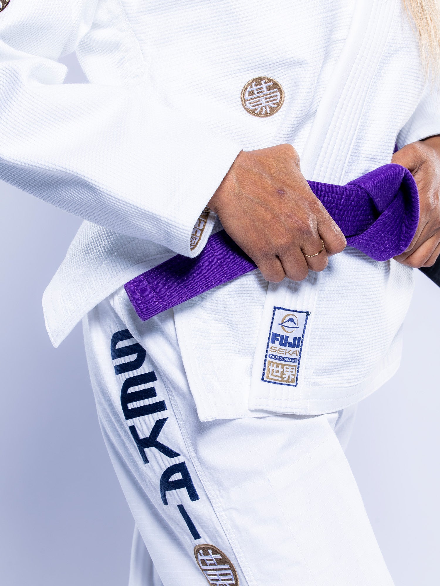 A person wears the Womens SEKAI 3.0 BJJ Gi White Navy with patches and ties a purple belt around their waist. The gi displays Japanese characters and the FUJI SEKAI logo on the jacket and pants.