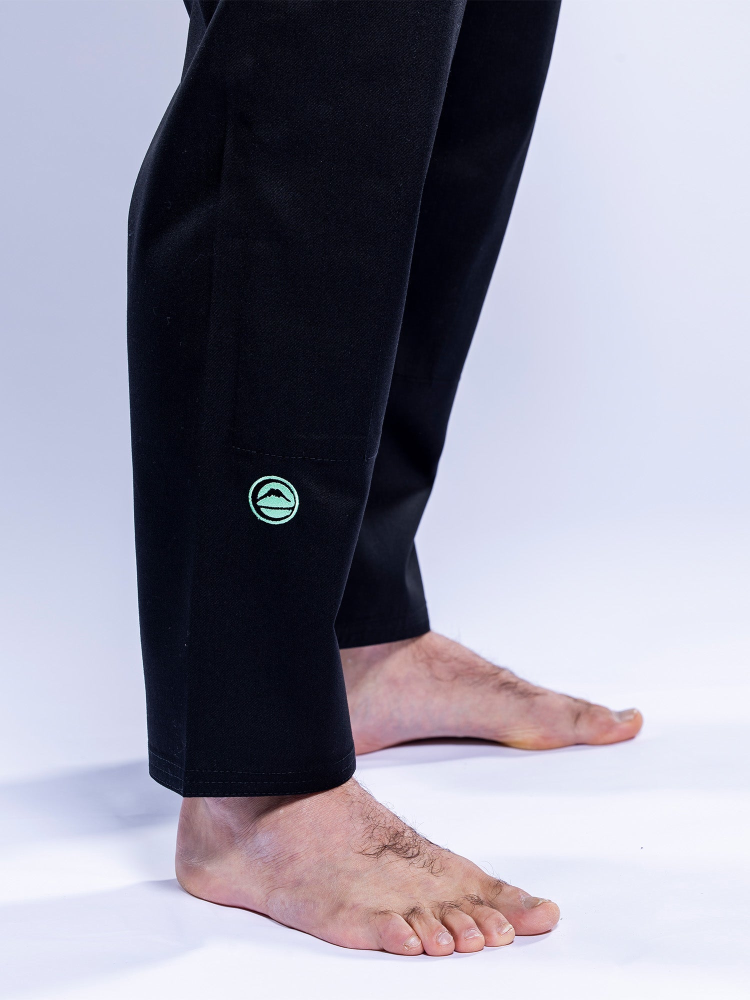 A person stands barefoot on a white surface, wearing the Baseline 2.0 BJJ Gi Black Mint pants, which feature a small green circular logo near the ankle. The image focuses on their lower legs and feet.