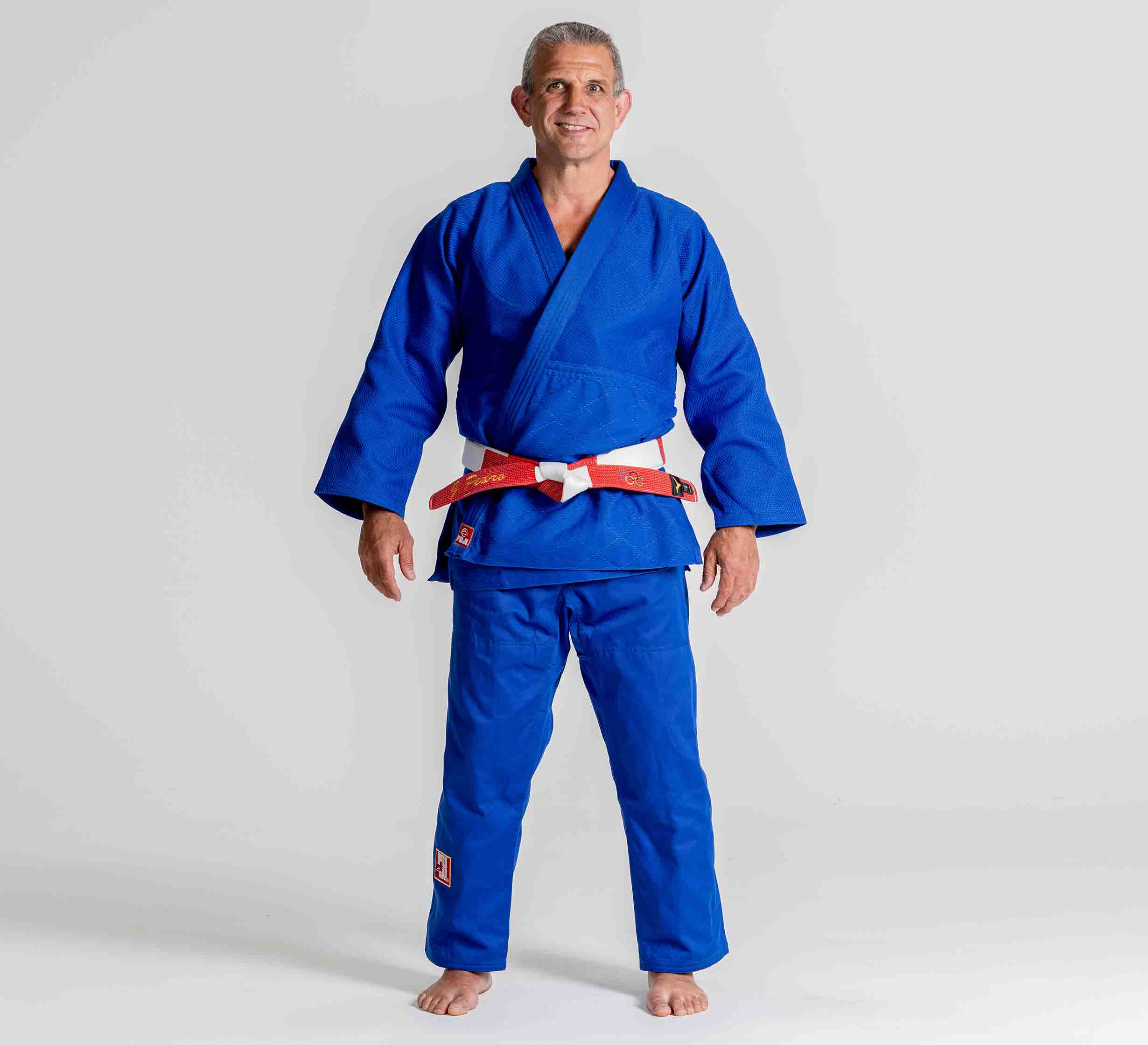 A person stands barefoot on a white background, smiling with arms relaxed at their sides while wearing the Euro Competition Judo Gi Blue and a red and white belt.