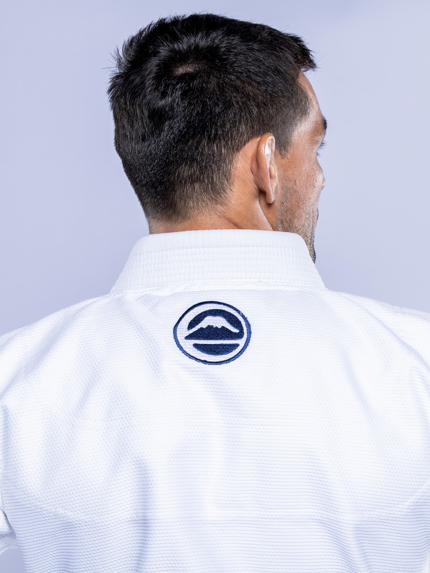 A man with short dark hair is seen from behind wearing the SEKAI 3.0 BJJ Gi White Navy, which features a circular black and white logo with two wave shapes on the back of the collar.