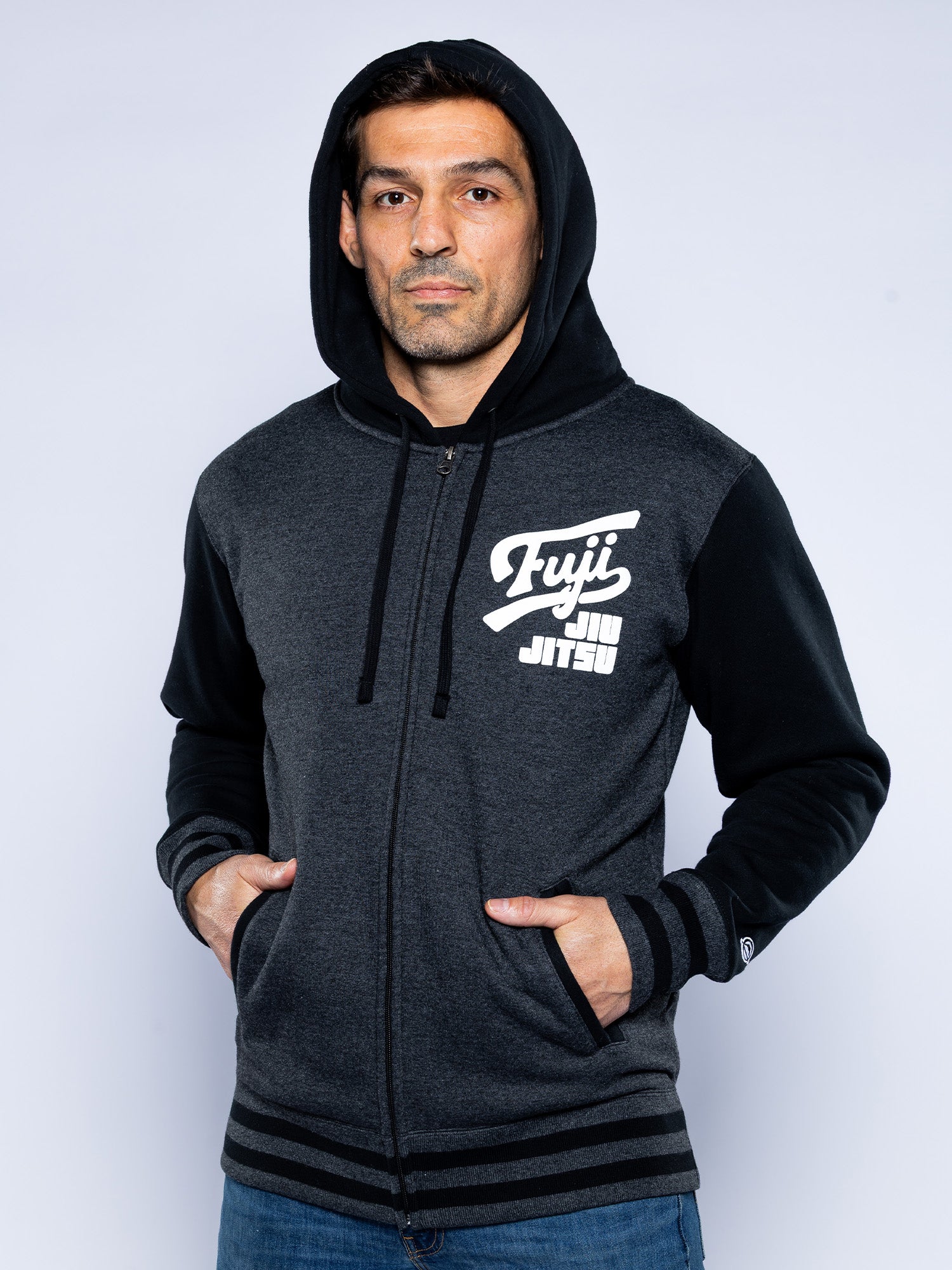 A man stands with his hands in his pockets against a plain light background, wearing the Jiu Jitsu Varsity Zip Hoodie Charcoal, a dark gray and black zip-up jacket with "Fuji Jiu Jitsu" printed on the left chest.