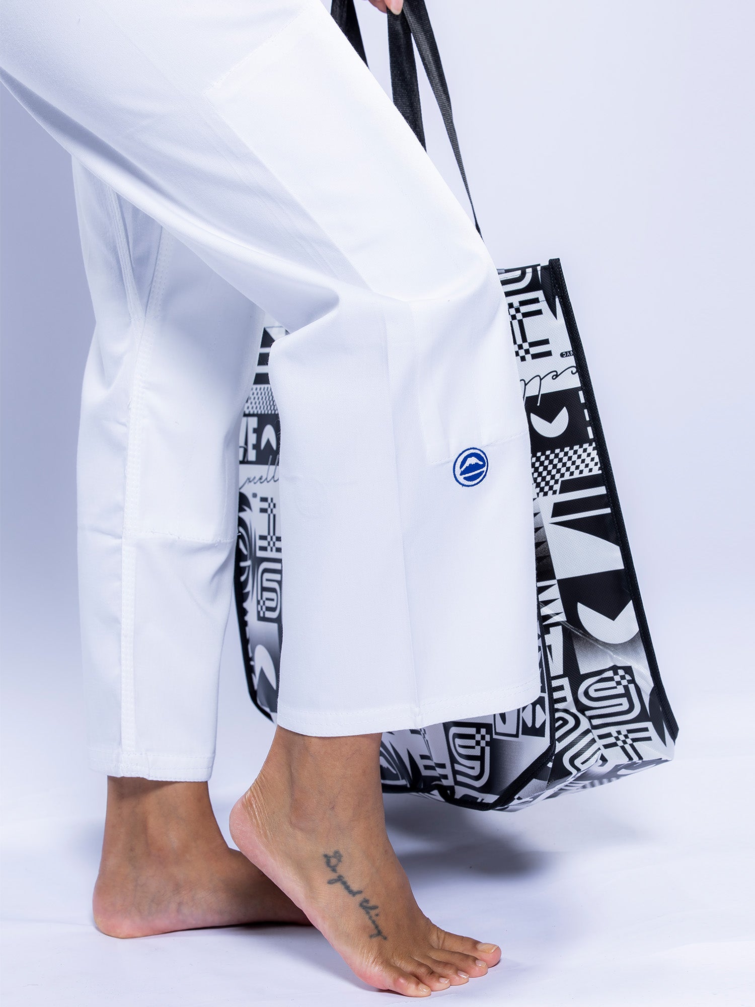 A person barefoot with a foot tattoo wears the Womens Baseline 2.0 BJJ Gi White Navy and carries a black and white patterned tote bag against a white background.