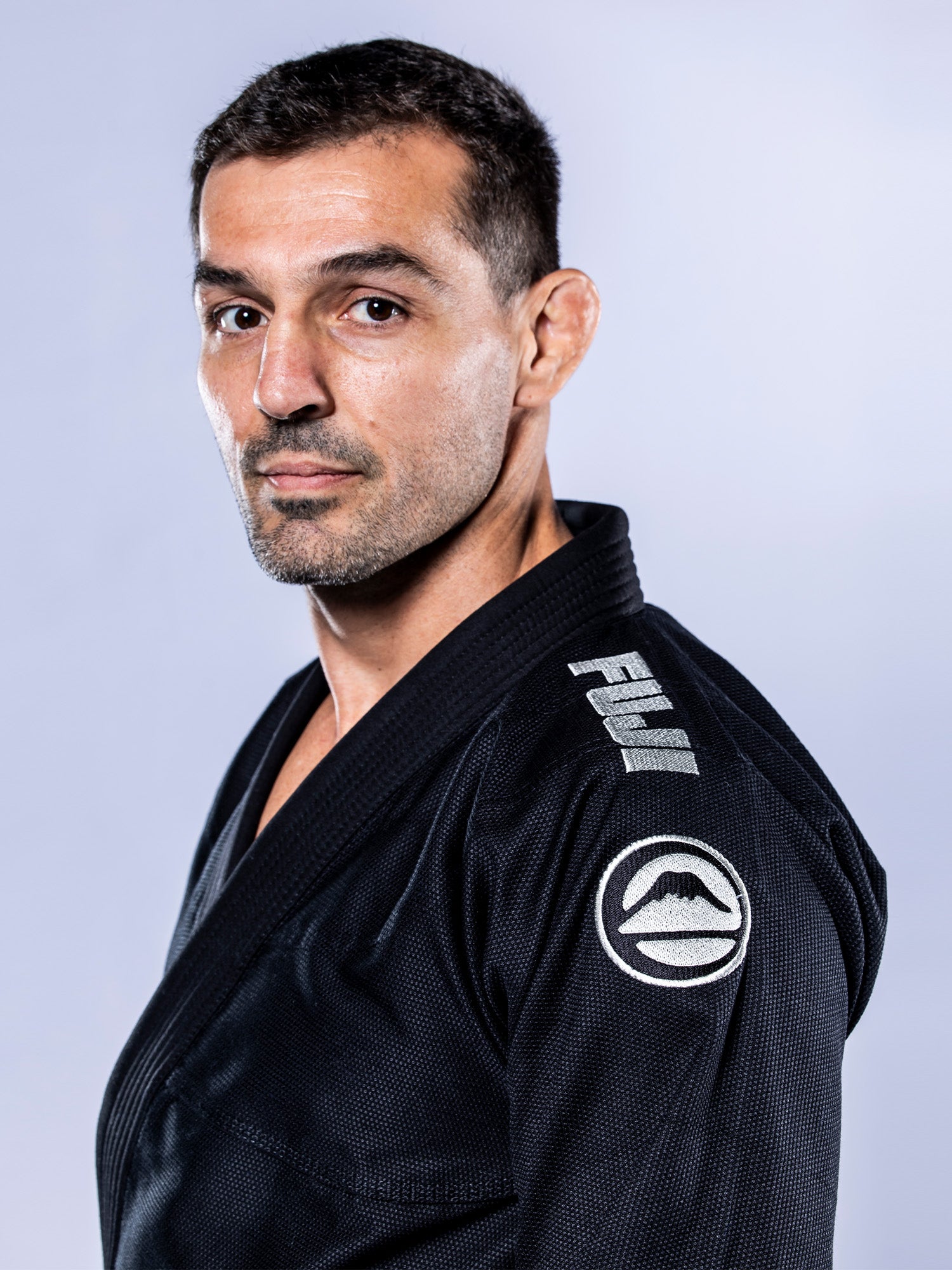 A man with short dark hair and light stubble wears the Baseline 2.0 BJJ Gi Black Grey by FUJI, featuring a mountain logo on the shoulder, as he poses against a plain light background.