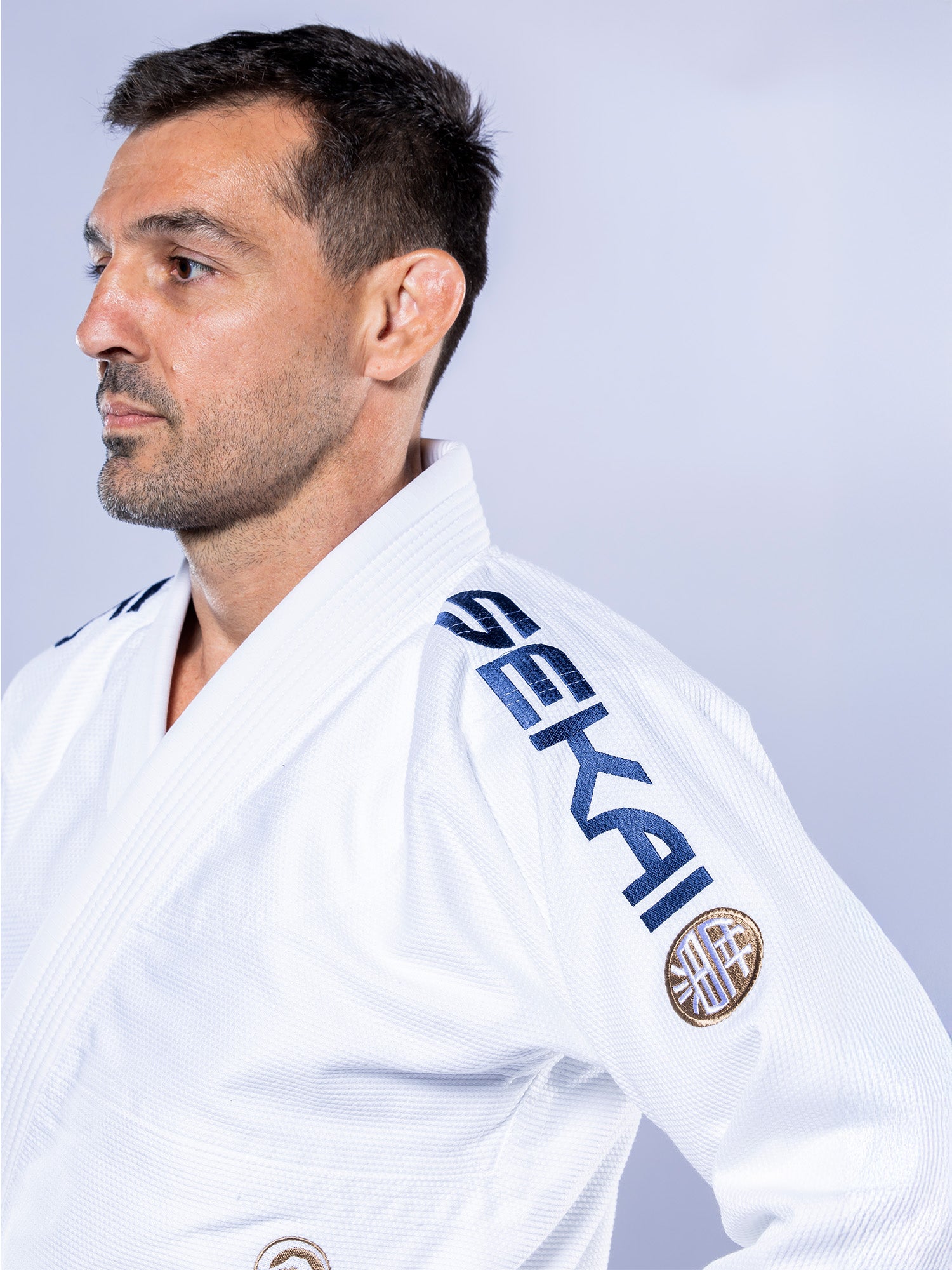 A man with short dark hair and a trimmed beard wears the white-navy SEKAI 3.0 BJJ Gi, embroidered with "SEKAI" on the shoulder, looking left against a plain light background.