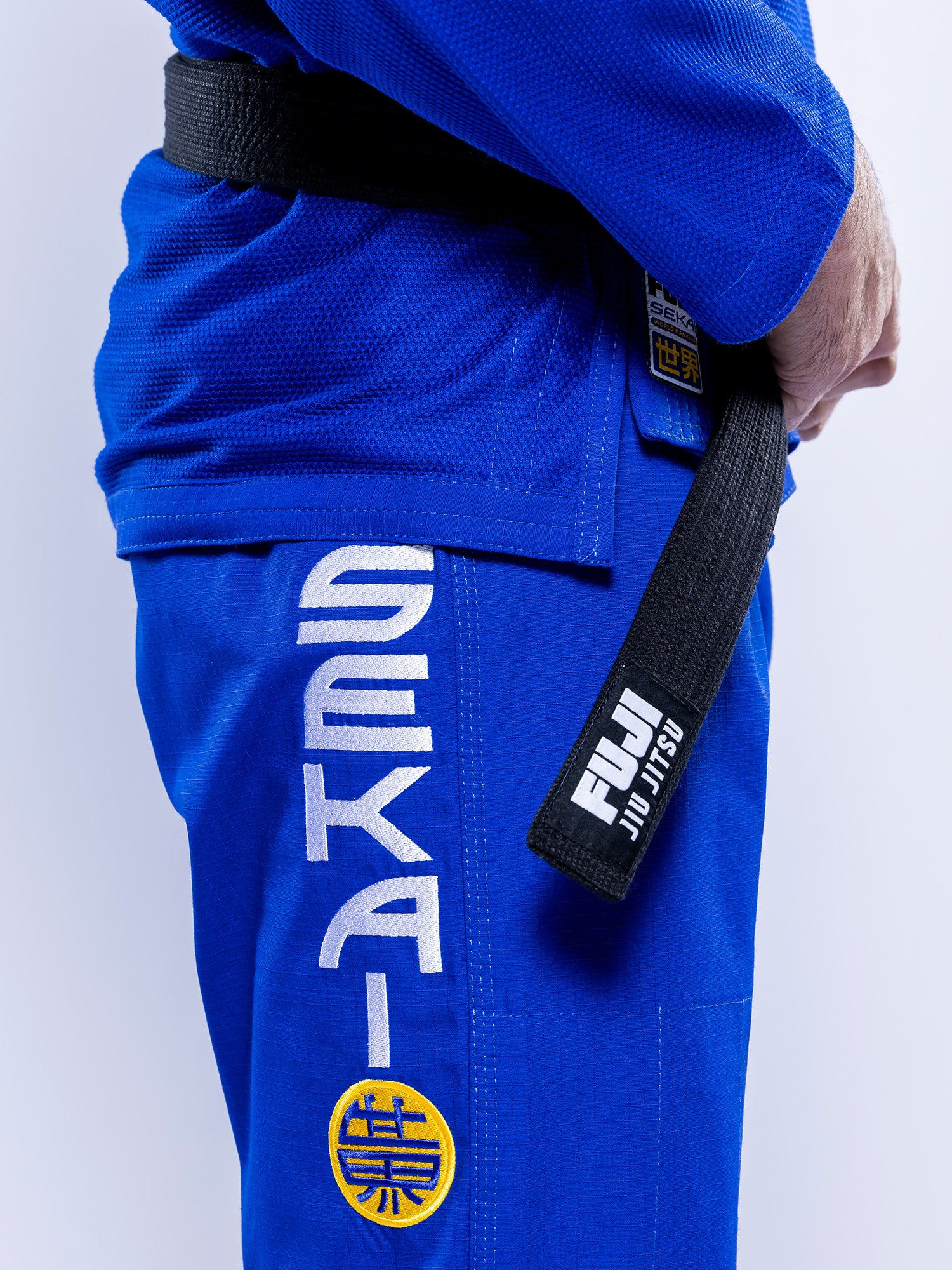 A person wearing the SEKAI 3.0 BJJ Gi Blue Yellow, featuring “SEKAI” and a yellow patch on the pants, holds a black Fuji Jiu Jitsu belt. The photo captures their torso and arm against a light background.