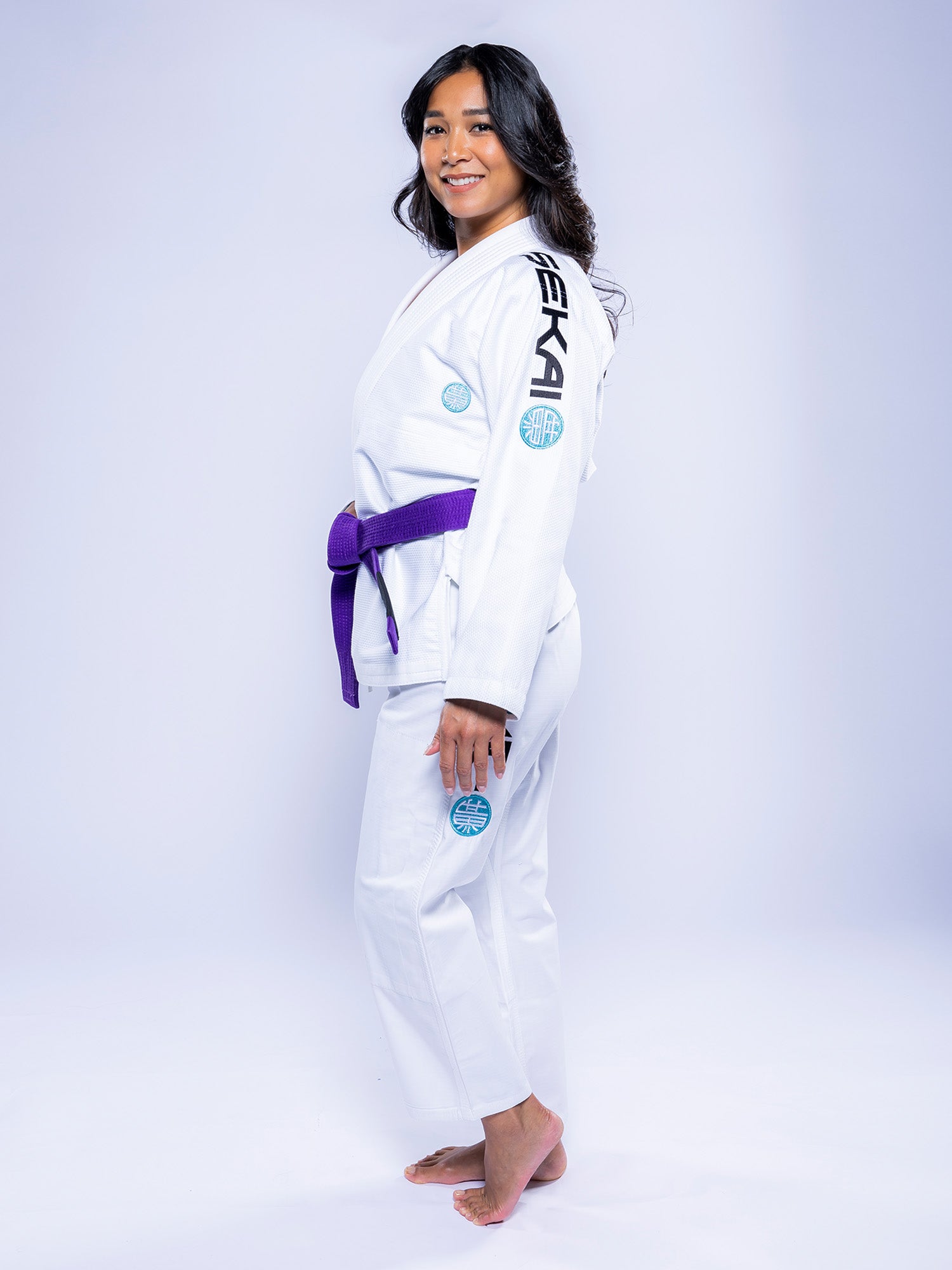A woman stands barefoot on a white background, smiling and facing sideways with her head turned toward the camera. She is wearing the Womens SEKAI 3.0 BJJ Gi White Teal, featuring a purple belt and black and blue patches.
