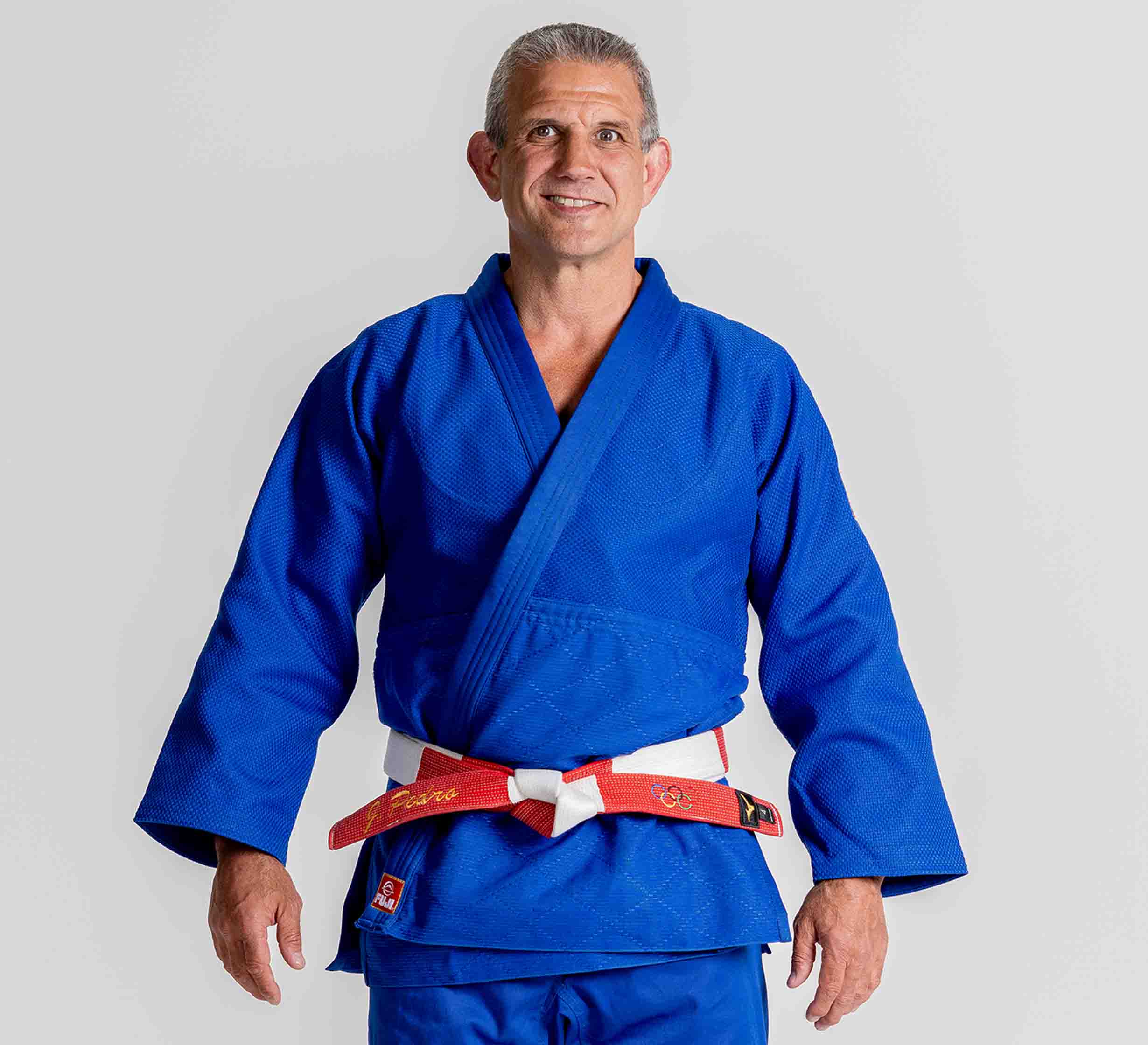 A man wearing the Euro Competition Judo Gi Blue and a red-and-white belt stands facing forward, smiling with his arms relaxed at his sides against a plain light background.
