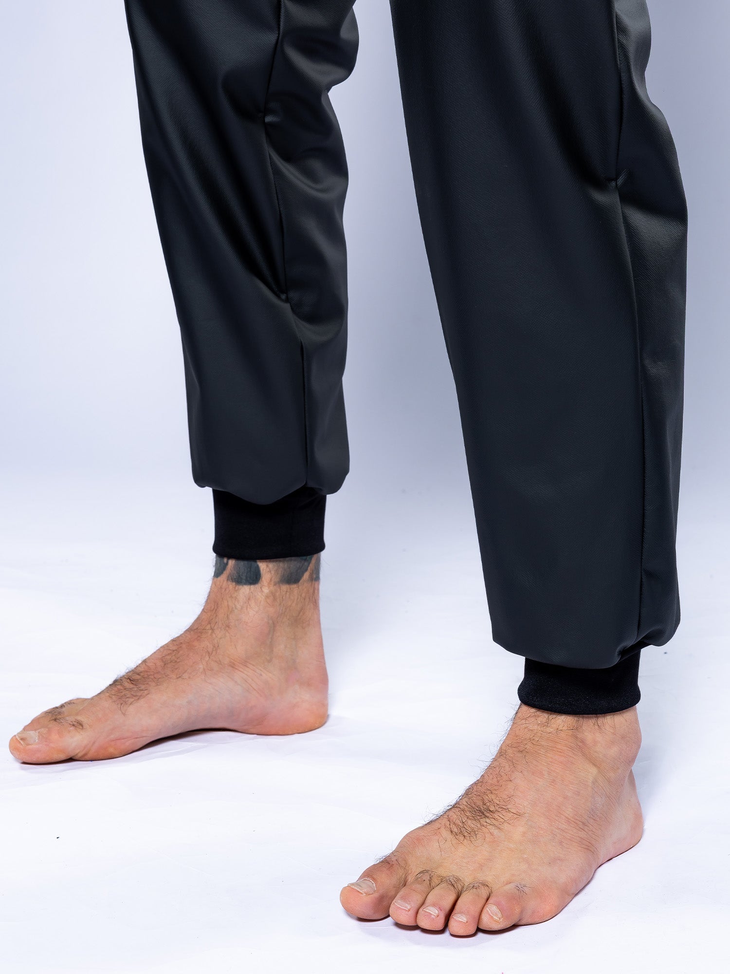 Close-up of a person’s bare feet and ankles wearing the Grappling Sauna Suit Black with cuffed pants. The background is plain and light-colored, and a dark tattoo is visible on their left ankle.