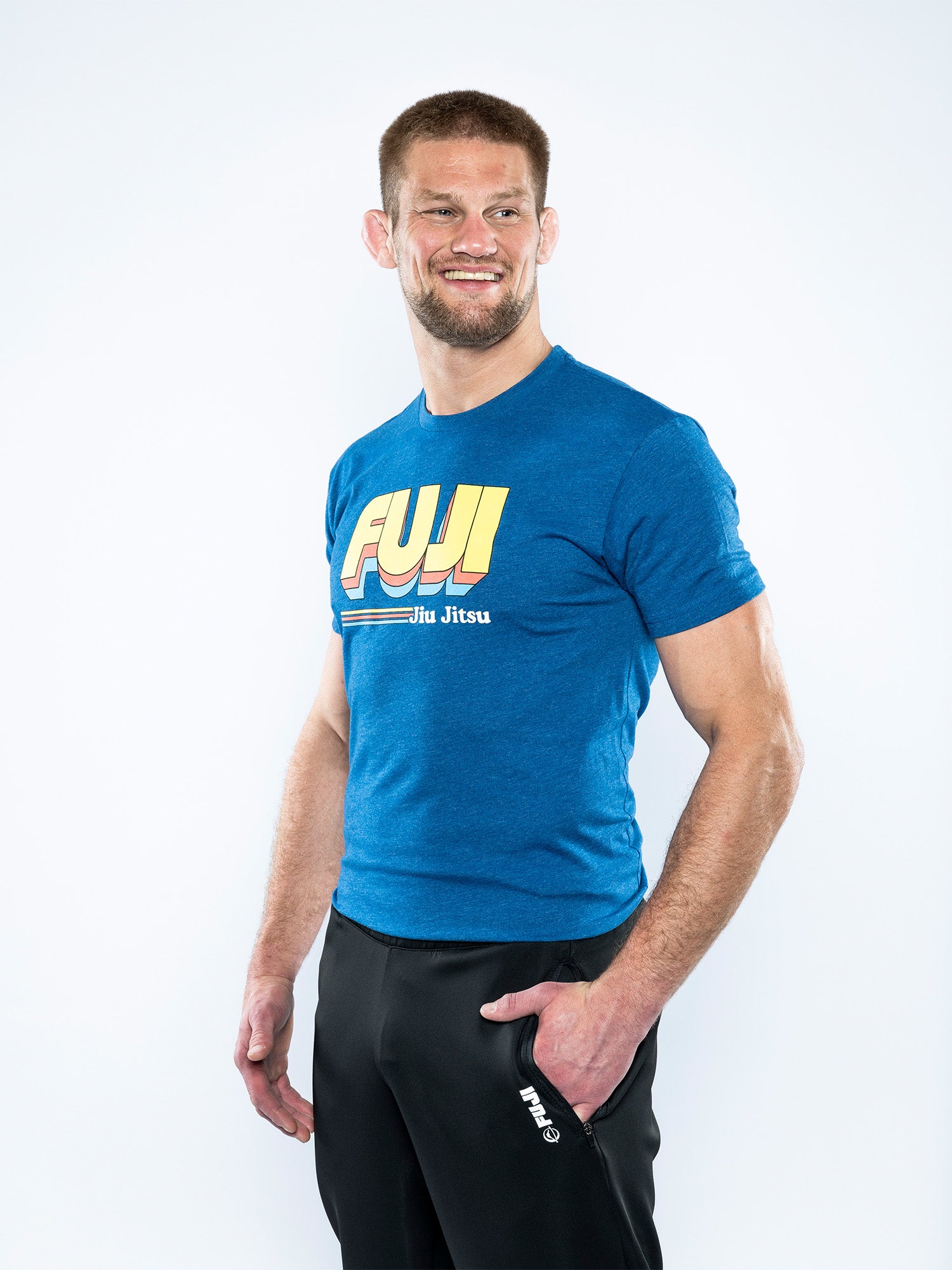 A man with short hair and a beard smiles against a white background, wearing the Vintage Jiu Jitsu T-Shirt Blue and black pants, with one hand in his pocket.