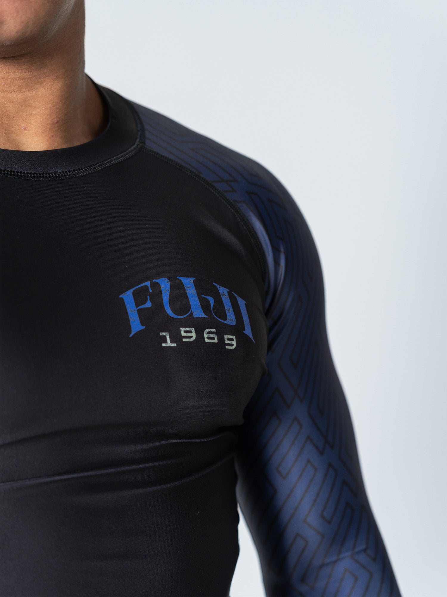 A person wearing the Oni Samurai Flex Lite Rashguard Blue, a black and blue long-sleeve athletic shirt with "FUJI 1969" on the chest. Only the upper torso and part of one arm are visible.