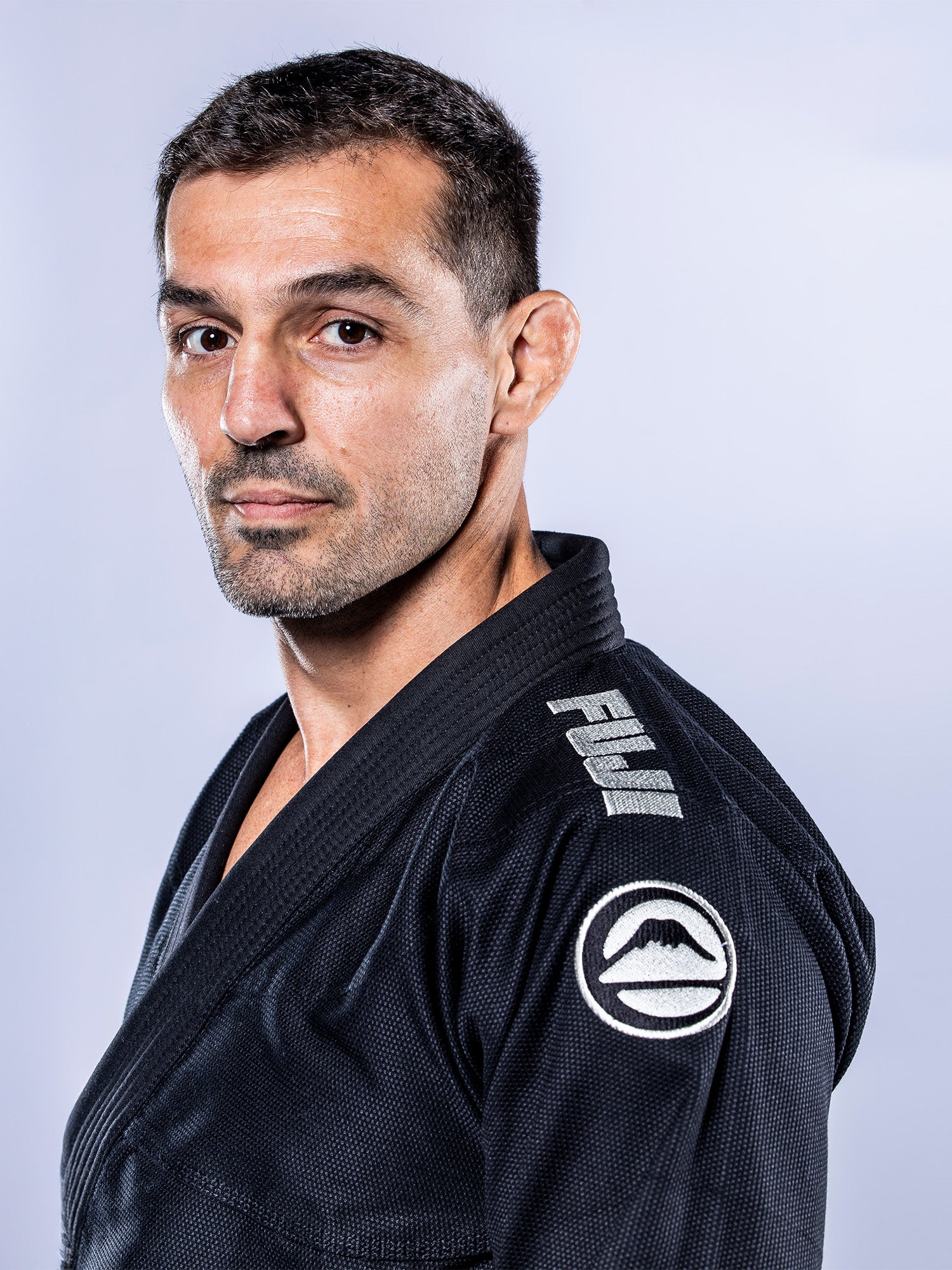 Man with short dark hair and trimmed beard wears the Baseline 2.0 BJJ Gi Black Grey by FUJI, featuring a logo on the collar and a sleeve patch, standing against a plain light background and looking at the camera.