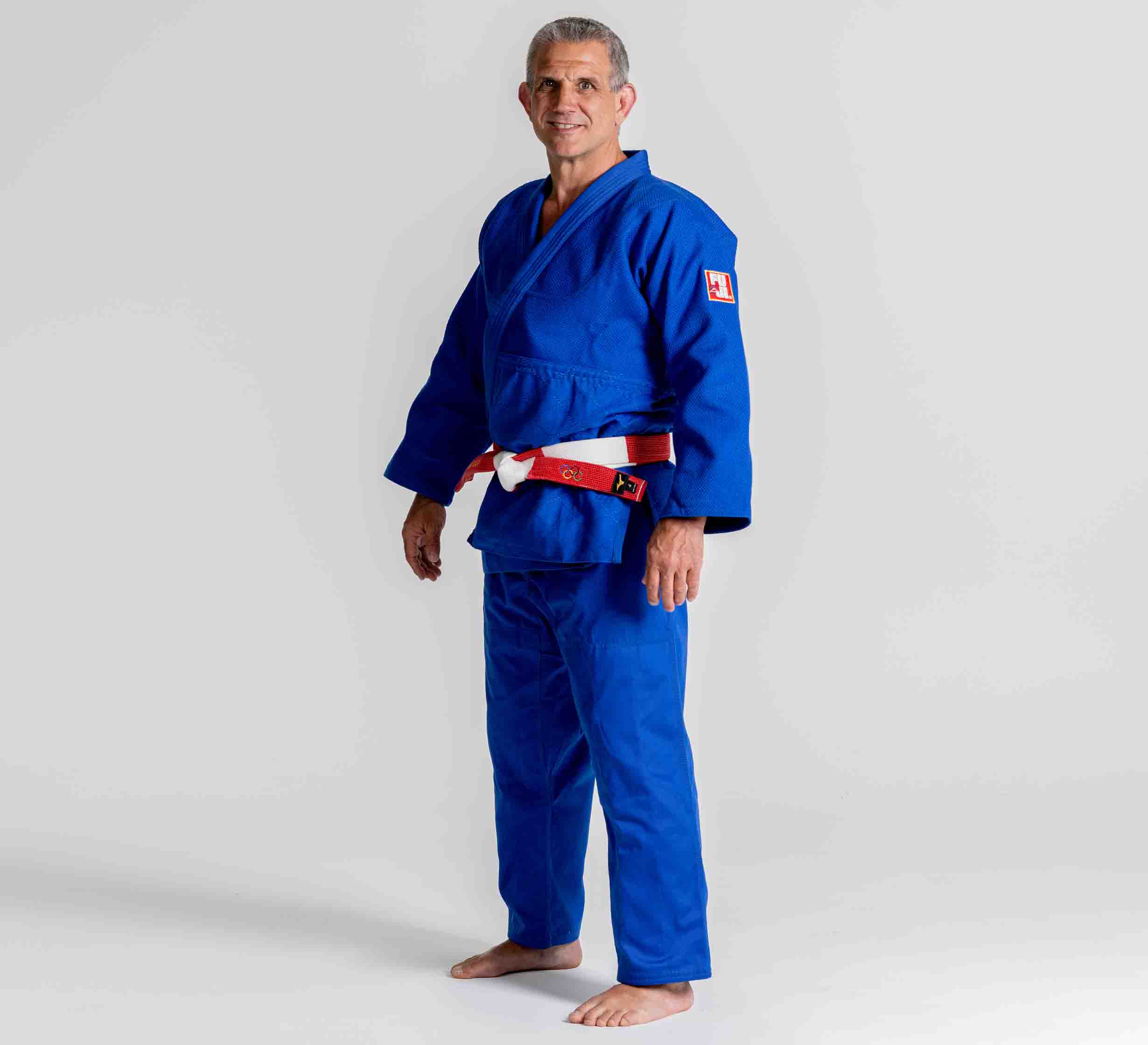 A smiling man stands barefoot on a plain background, wearing the Euro Competition Judo Gi Blue and a red and white belt. His stance is relaxed, with his arms by his sides.