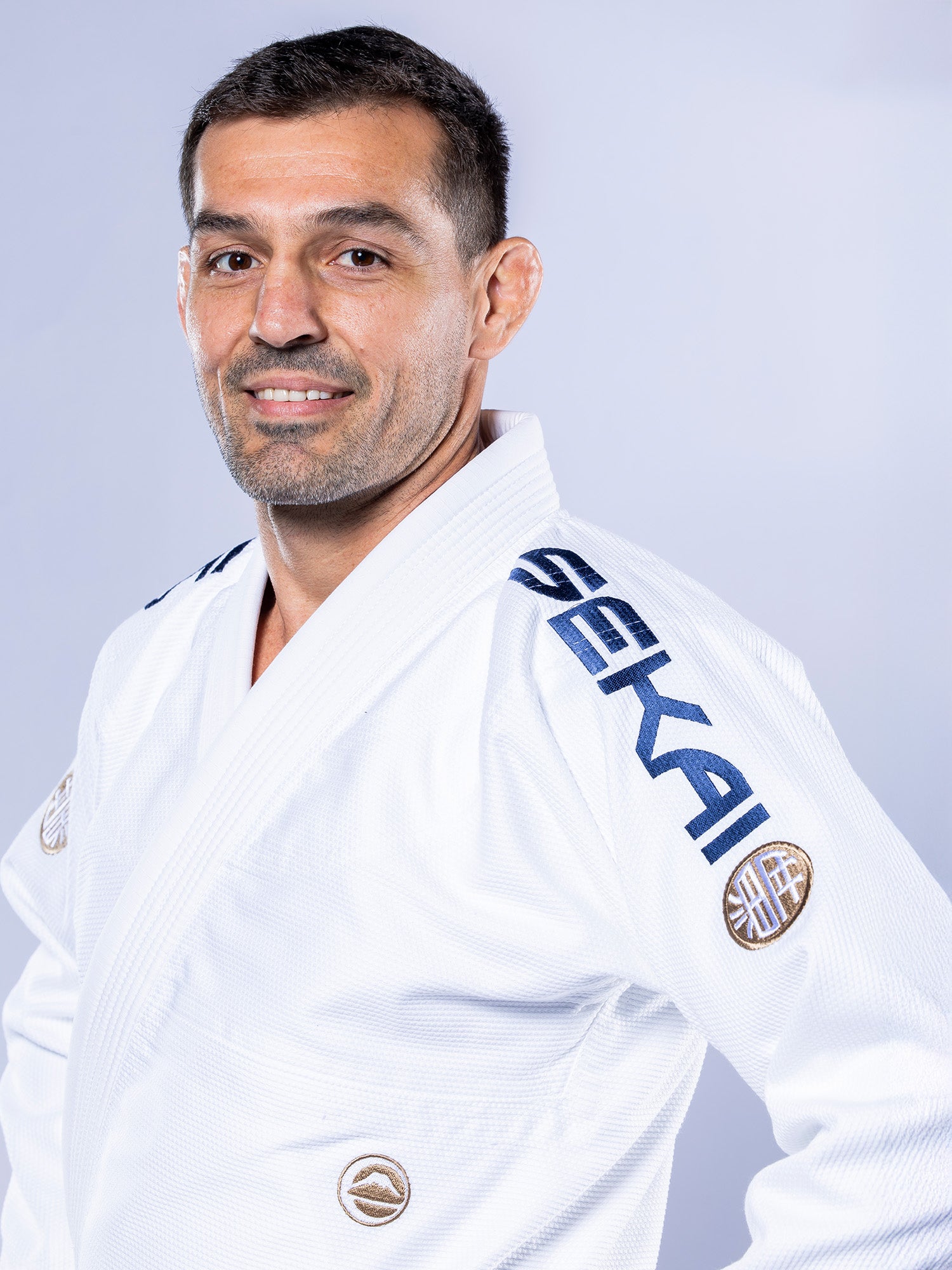 A man wears the SEKAI 3.0 BJJ Gi White Navy, featuring "SEKAI" on the shoulder, and stands against a plain light background, smiling slightly at the camera.