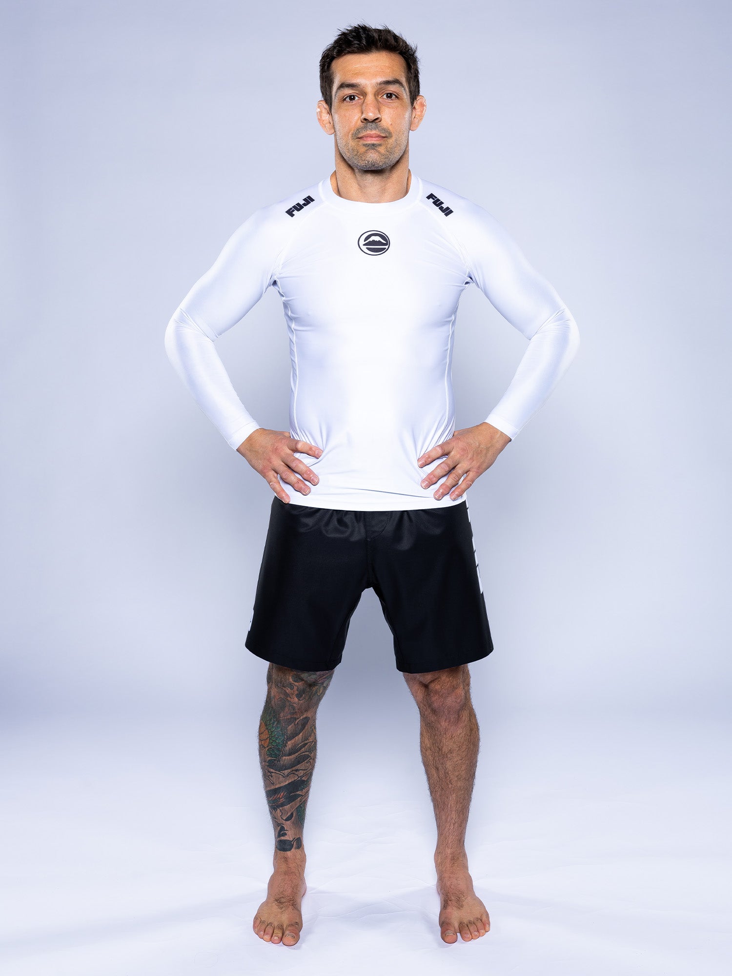 A man stands barefoot on a white background, wearing the Baseline 2.0 Long Sleeve Rashguard White with black logos on the shoulders and plain black shorts. He has tattoos on his left leg and his hands rest on his hips.